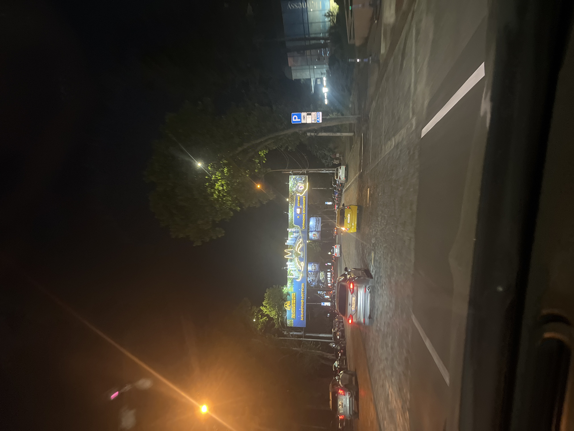 The image depicts a night-time street scene in Tbilisi, Georgia. It appears to be a view from inside a vehicle looking forward. A street with multiple lanes of traffic is visible, with several cars driving and parked. A large billboard-like structure with images and text is suspended over the road, featuring what looks like an advertisement for "Ambassador Island Batumi" and "Expanding Boundaries". Yellow streetlights illuminate the scene, casting a warm glow. Trees line the street, and in the background, a building with illuminated windows can be seen. A blue parking sign is also visible on a pole. The road surface appears to be paved with cobblestones in some areas and asphalt in others. The overall atmosphere is that of a typical urban street at night, with traffic and commercial signage.