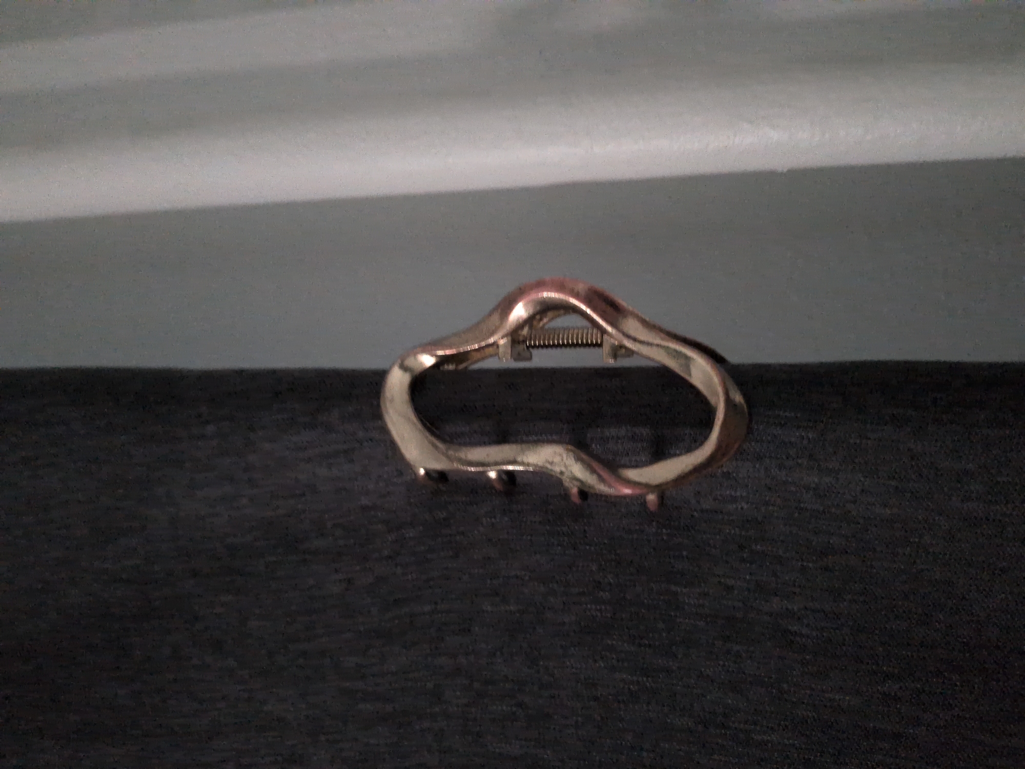 In Unknown, Unknown, a vertical close-up photograph displays a metallic clamping device affixed to dark, textured fabric, with lighter drapery in the background. The left half of the image features a tightly woven, dark charcoal-gray fabric with a subtle mottled pattern. Centrally positioned, the irregular, curvilinear metal object possesses a polished, warm golden-bronze finish, exhibiting reflections from an unseen light source. On its left side, three small, rounded metallic studs are visible, pressing into the dark fabric. The right side of the object reveals a functional spring mechanism with a visible threaded screw component, indicative of a clamping or tensioning design. To the right of the metallic device, the background consists of a lighter, neutral gray fabric, displaying faint vertical folds or creases characteristic of a curtain. The scene is evenly lit, with specular highlights on the metal.