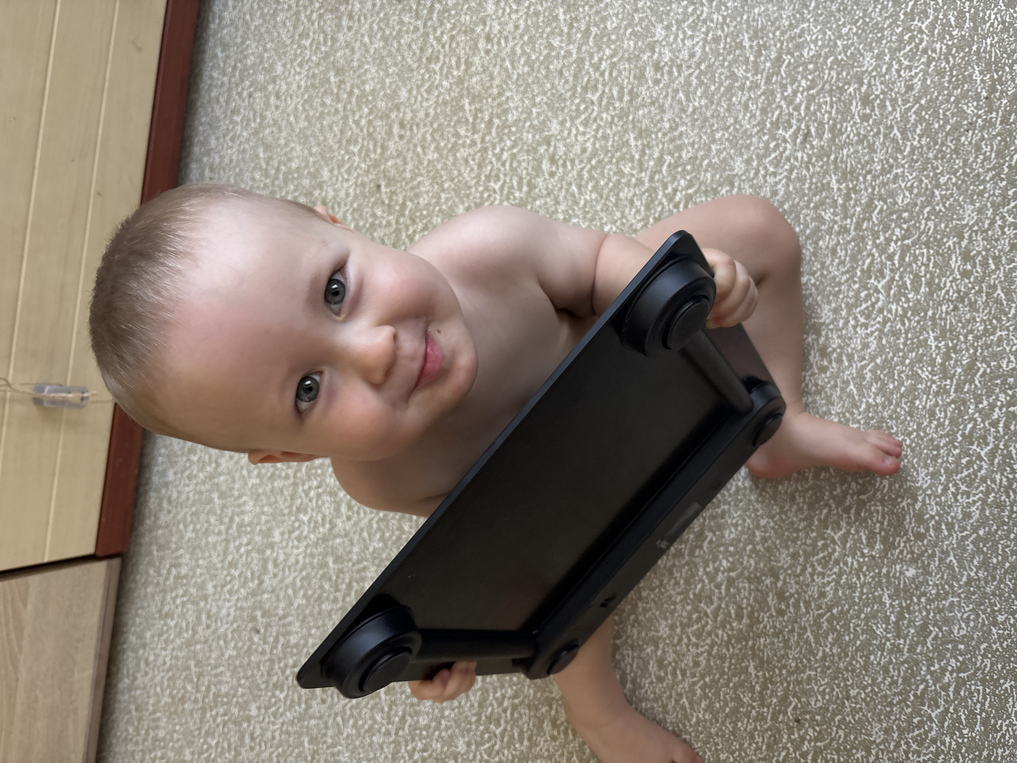 A baby is sitting on a carpeted floor, holding a black digital scale. The baby has fair skin, light brown hair, and blue eyes. The baby is looking up at the camera with a slight smile. The scale is rectangular and black, with round feet at each corner. The baby is partially undressed, wearing only a diaper. The background is a light-colored carpet with a textured pattern. To the left of the baby, there is a wooden cabinet or furniture piece. The lighting suggests it is daytime, possibly indoors. The overall scene is intimate and captures a candid moment of a baby interacting with an object. There is no visible text in the image.