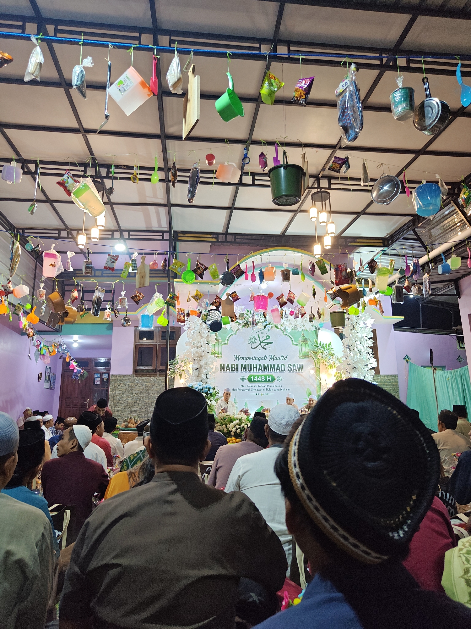 This image depicts an indoor gathering, likely a religious event, held in Cermee, Indonesia. The space appears to be a decorated hall with a ceiling adorned with various household items hanging from strings, including buckets, pots, pans, cutting boards, and plastic containers. In the background, a stage is set up with white floral decorations and a banner that reads "Memperingati Maulid Nabi Muhammad SAW" (Commemorating the Birthday of Prophet Muhammad SAW) and "1448 H". Several men, mostly wearing peci caps or other traditional headwear, are seated facing the stage, suggesting an audience. Some individuals are dressed in formal attire, while others wear more casual clothing. The lighting appears to be artificial, with hanging lamps visible in the ceiling. The overall atmosphere suggests a community event or celebration.