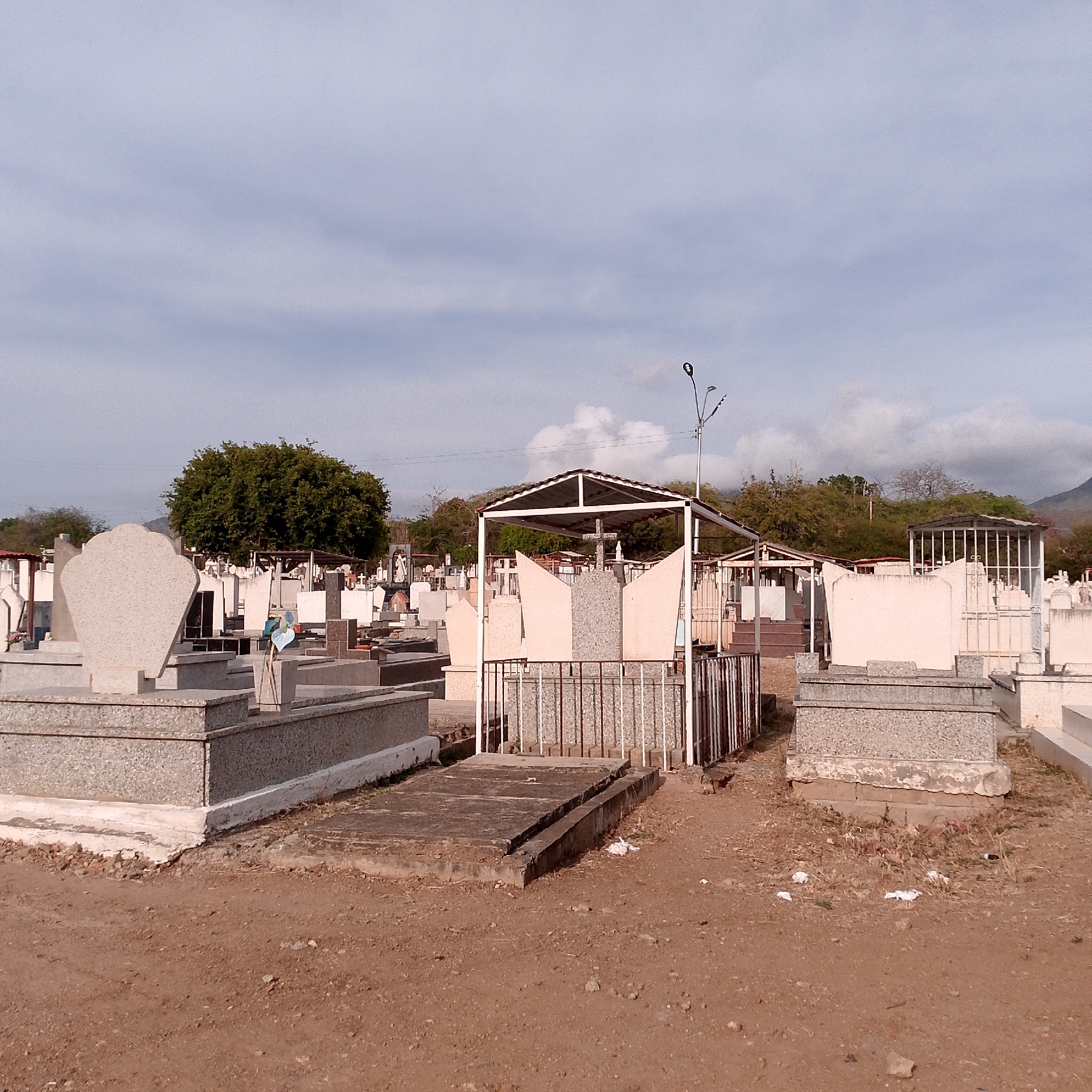 The image depicts a cemetery in Altagracia, Venezuela, under an overcast sky. Numerous gravesites are visible, varying in design and material. Some are simple concrete slabs flush with the ground, while others are more elaborate structures with headstones, crosses, and protective coverings. A few gravesites have metal fences or railings. The ground is primarily dirt, with scattered debris and dry vegetation. In the background, there are trees and a hill. A tall light pole stands amidst the gravesites.