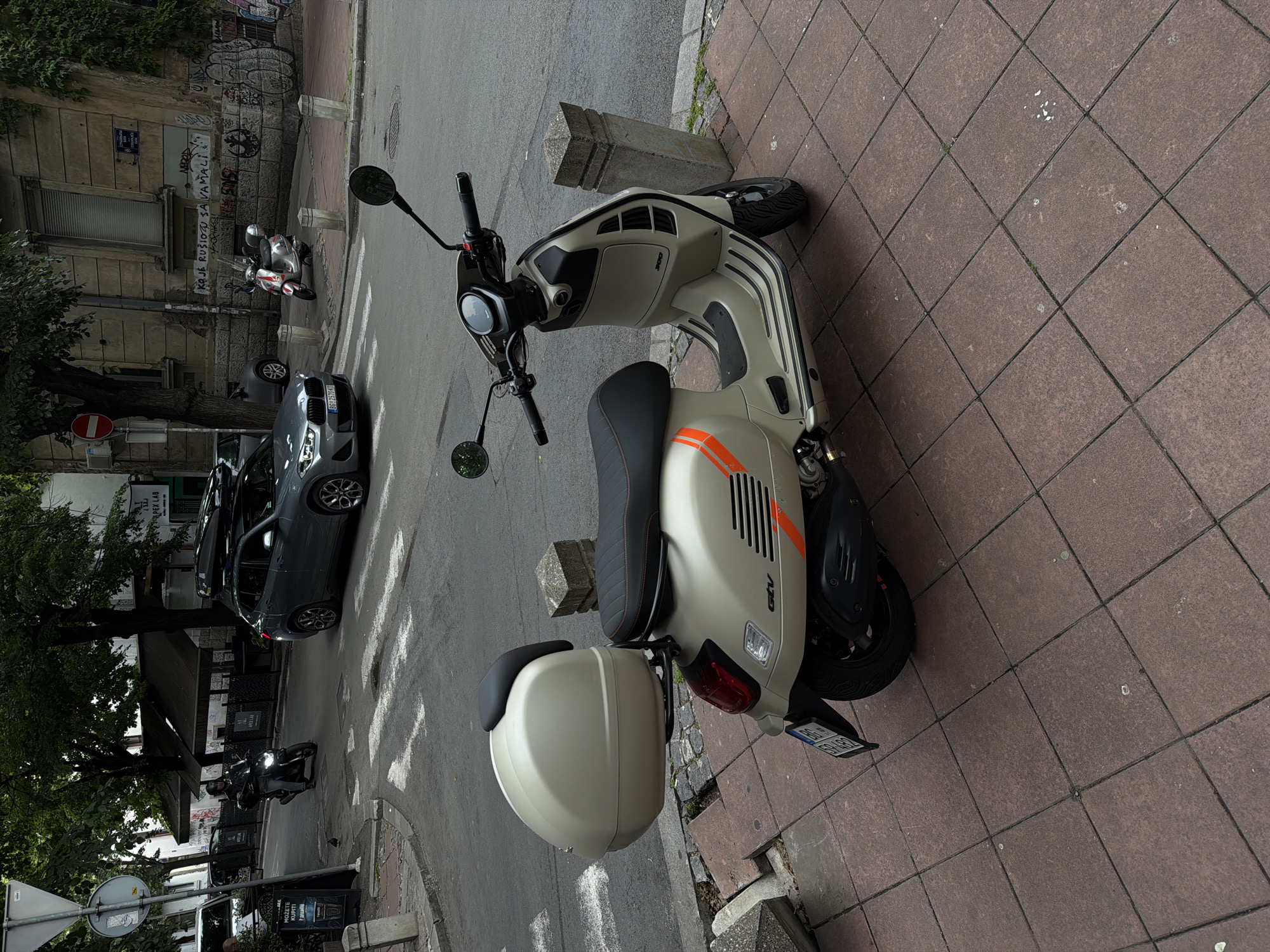 The image captures a light beige Vespa GTV 300 scooter parked diagonally on a brick-paved sidewalk. The scooter faces into the street. It has black and orange stripes on the side and a storage box behind the seat. The license plate reads "BG 2575-KZ." In the background, there's a grey car, a passing motorcyclist, and part of a building covered in graffiti. Signage for "ARCALFA APEX LAB" and "PEX LAB" is visible. The area is identified as being in Belgrade, Serbia, based on the license plate and the Cyrillic graffiti on the building. The street appears to be a mix of commercial and residential buildings. A sign with "KOJE JE RUSIOZA SA VAMALI" is also visible.