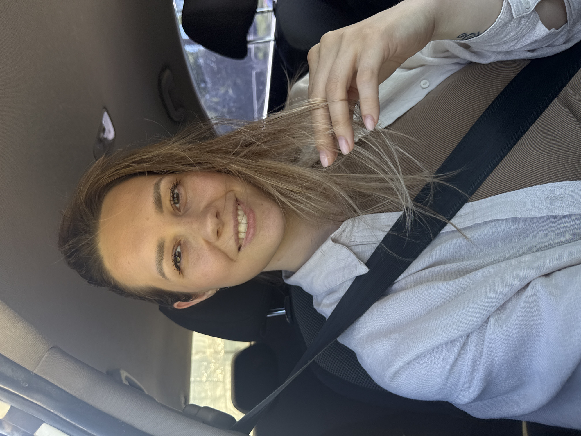 A young woman with shoulder-length blonde hair is smiling and seated in a car, wearing a brown top and a white button-down shirt. A black seatbelt is fastened across her chest. Her left hand is near her hair. The interior of the car and the car window, showing an indistinct outdoor scene, are visible. It is not possible to determine if the location is Chaikovsky, Russia from the image.