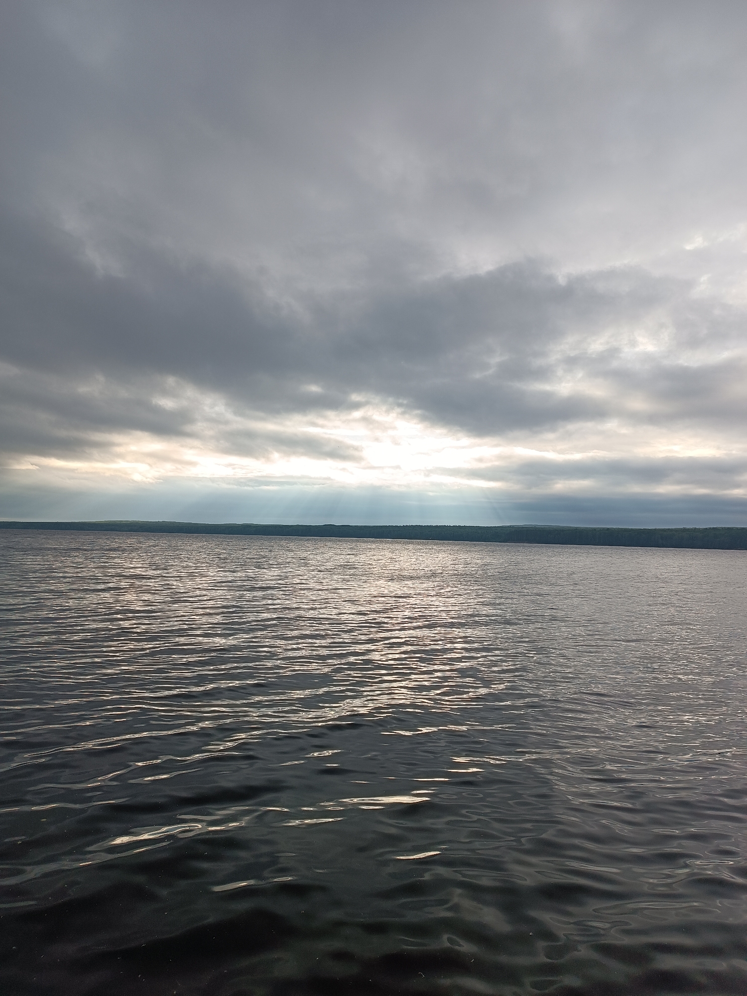 The image depicts a wide expanse of dark water with rippling waves, reflecting the light from the sky above. The sky is overcast with heavy grey clouds, but a break in the clouds allows rays of sunlight to stream down, illuminating a portion of the water and creating a luminous effect. In the distance, a dark, tree-covered shoreline is visible, forming a horizon line against the sky. The scene appears to be a natural landscape, likely a large body of water such as a river or lake. This location is identified as being in the city of Chaikovsky, Russia. There are no people, objects, or actions present in the image.