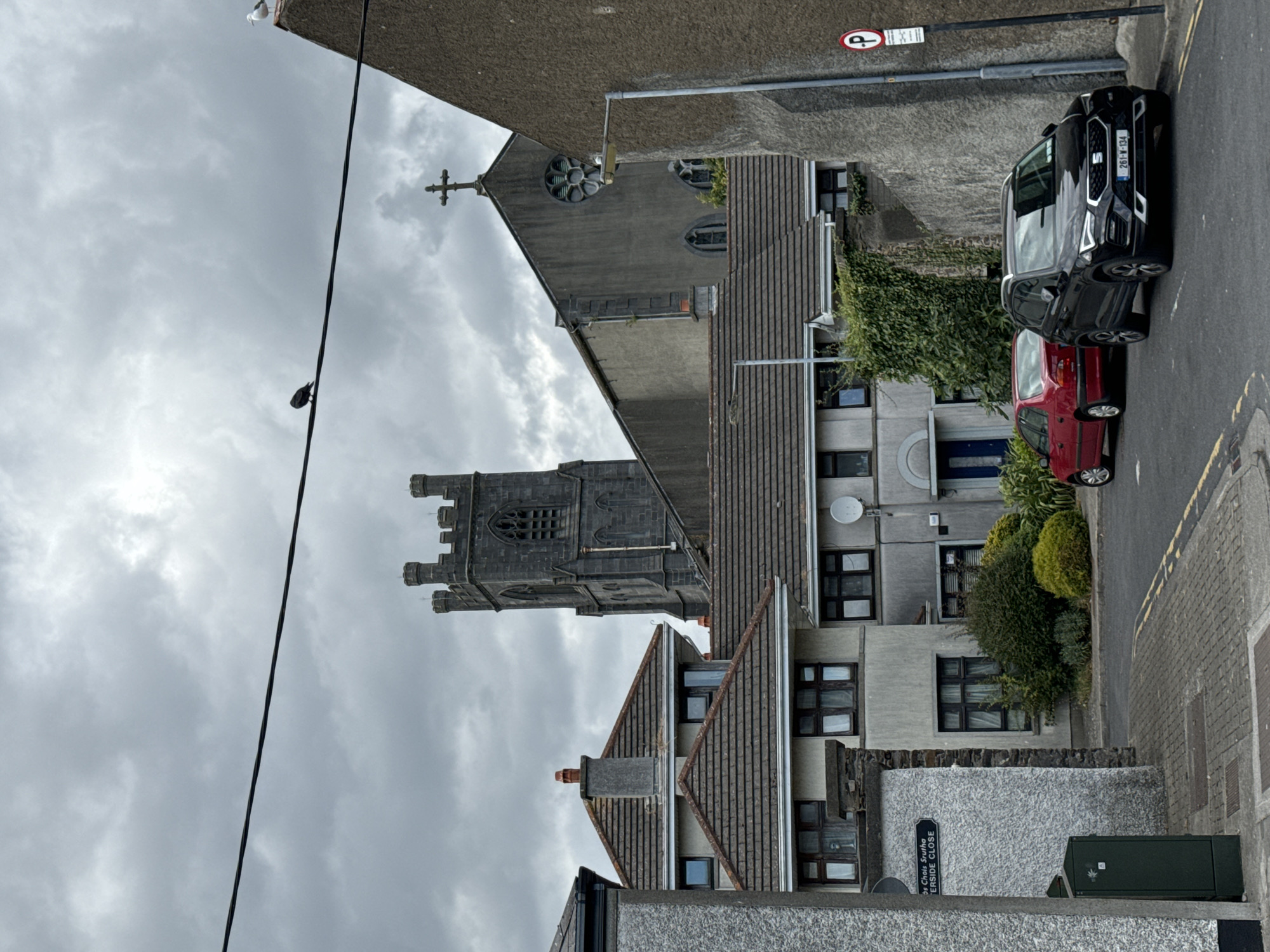 This is an exterior, eye-level, medium shot of a street in Waterford, Ireland, on a cloudy day. A dark colored SUV and a red sedan are parked on the right side of the street. Behind the cars, a stone wall covered in ivy runs along the street. A lamppost with a "P" sign is mounted on the wall. Further back, a church with a tall steeple and stained glass windows dominates the skyline. Houses with tiled roofs and windows line the street on the left. A street sign reads "58 CHOIS SRUTHA RIVERSIDE CLOSE". A bird is perched on a power line that stretches across the frame.