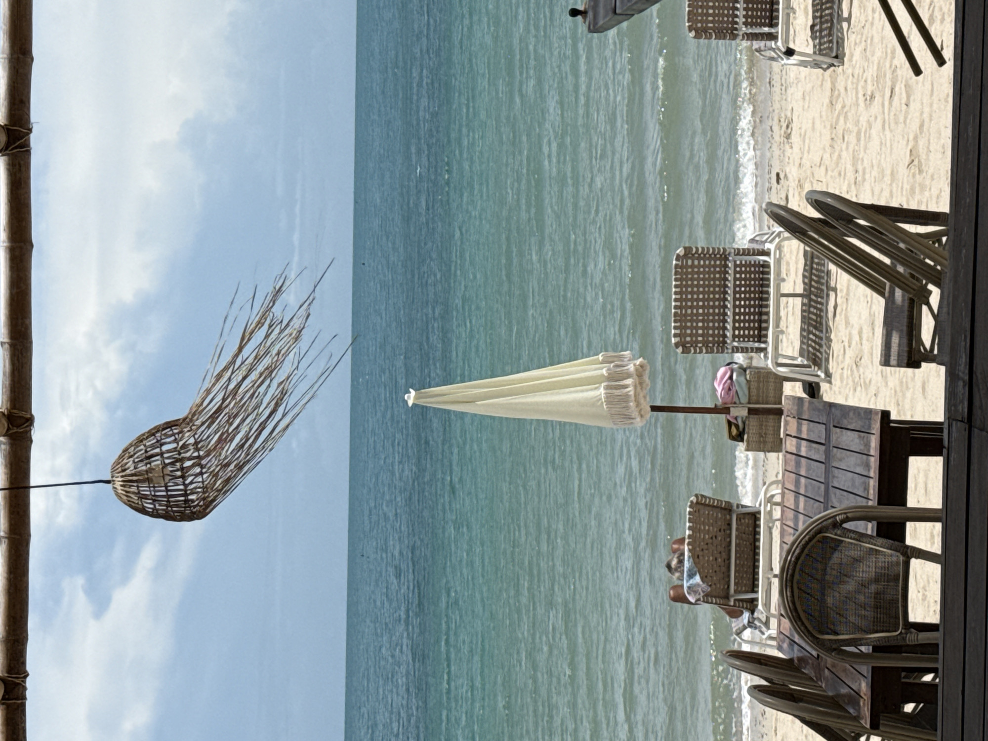 This image depicts a beach scene in Baan Tai, Thailand, characterized by a tranquil atmosphere. In the foreground, a wooden table with woven chairs is set on the sandy beach, facing the turquoise ocean. A white umbrella with fringed edges stands near the table. In the middle ground, two lounge chairs are occupied by a person sunbathing on one and a basket with items on the other. The ocean stretches to the horizon under a partly cloudy sky. A decorative woven lampshade hangs from a bamboo structure in the upper portion of the frame, swaying gently in the breeze.