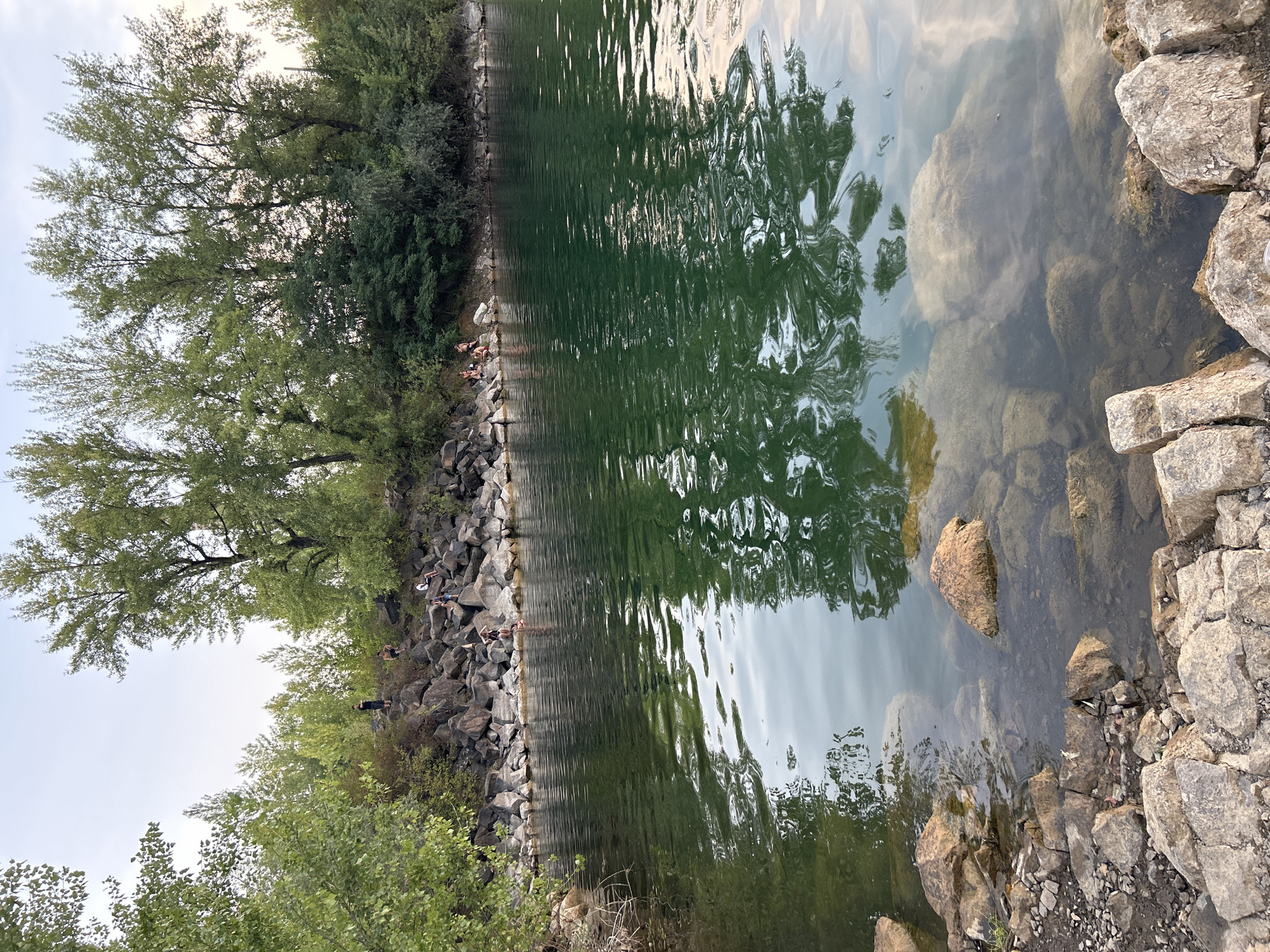 A group of people are seen relaxing by the water on a rocky shore, with lush green trees forming a backdrop against a pale sky. The water, a deep green, reflects the trees and the sky. Some individuals are seated on the rocks, while others appear to be standing or walking along the shoreline. The scene suggests a leisure activity in a natural setting. Without additional information or context, it is not possible to definitively place this image in the city of Uzhhorod, Ukraine, or any other specific location.