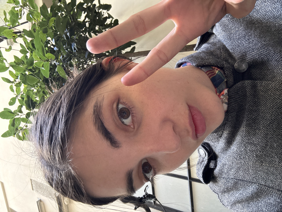 A close-up portrait captures a female individual with dark hair, brown eyes, and prominent eyebrows, looking directly into the camera. Her right hand is raised beside her face, forming a 'V' sign with the index and middle fingers extended. She wears a grey herringbone-patterned jacket, partially unbuttoned to reveal a blue, red, and white plaid shirt collar beneath. The background, illuminated by natural light, features a light-colored building facade with closed shutters visible on the upper left. To the upper right, green foliage from a tree or large plant is present. A dark, grid-like structure, possibly part of a window frame, is visible on the lower left. The architectural elements are characteristic of urban buildings in Paris, France, suggesting the scene takes place outdoors, likely within a courtyard.
