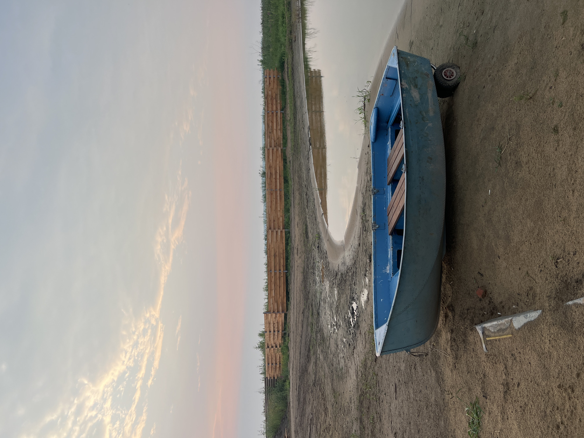 A weathered, blue rowboat with wooden seats rests on sandy ground near the edge of a calm body of water. The boat is equipped with two small wheels at its stern, suggesting it can be easily transported. A rustic wooden fence stretches across the background, separating the sandy shore from a grassy field and a line of distant trees under a pale, cloudy sky. The sky exhibits soft hues of pink and orange, indicative of either sunrise or sunset. The environment appears rural and tranquil, with no people visible in the scene. The overall impression is one of peaceful solitude in a natural setting.