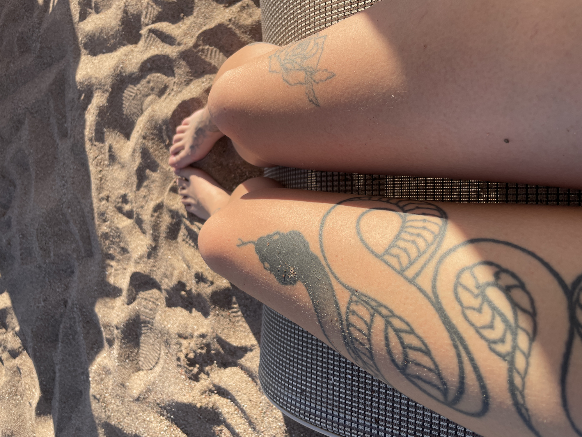 This is a high-angle, medium shot of a person's legs resting on a beach chair, with sand visible in the background. The person's left leg, closest to the camera, features a large black tattoo of a snake with detailed scales and a head. The right leg shows a smaller tattoo of a floral design. The sand is light brown and textured, with visible footprints and shadows cast by the sun, suggesting it is late afternoon or early morning. The person's feet are visible in the upper portion of the frame, resting on the sand. The scene appears to be on a beach in Rafailovići, Montenegro, characterized by the sandy terrain and the bright sunlight.