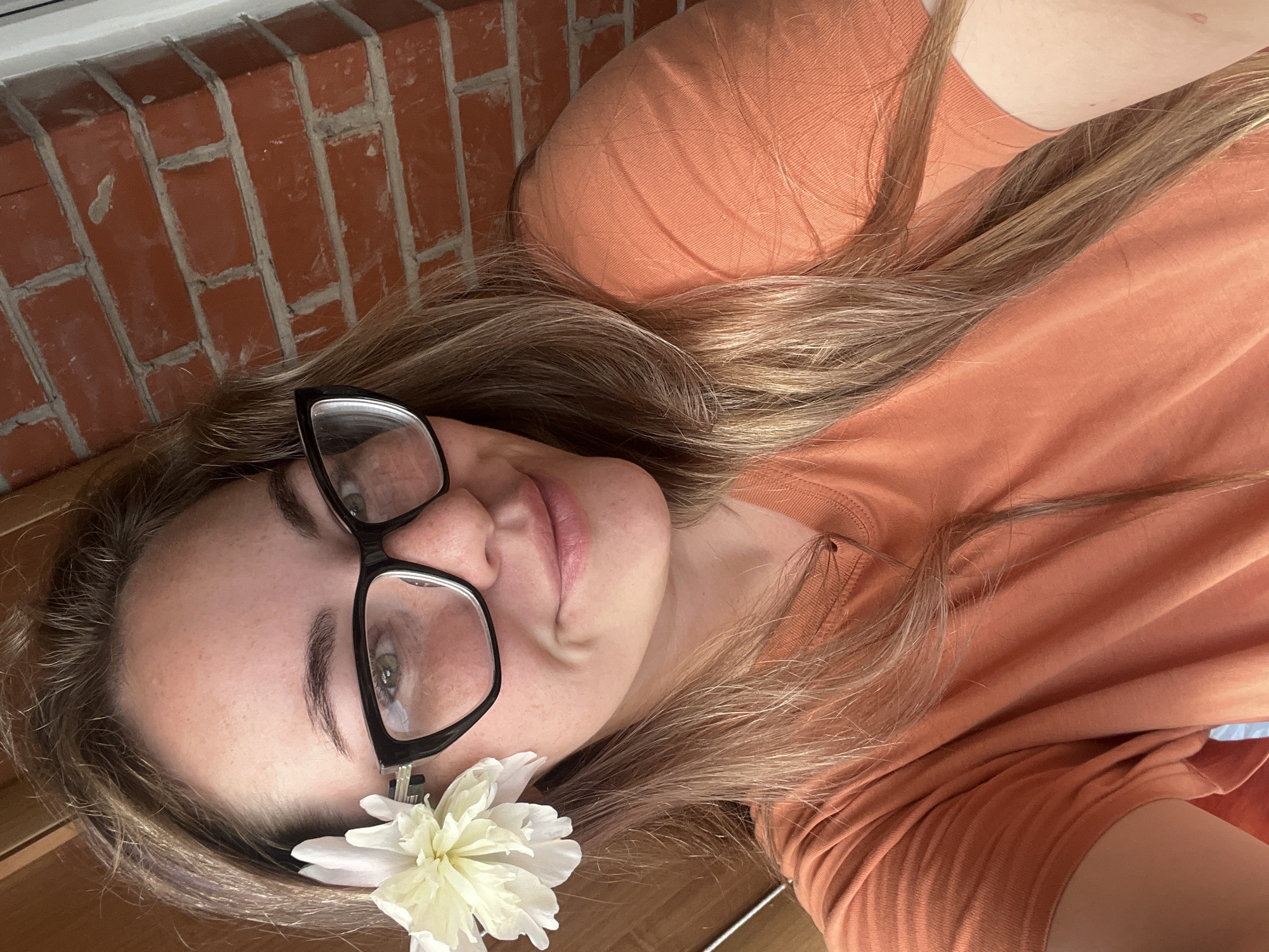 The image is a close-up, selfie-style shot of a woman with long, blonde hair and glasses. She has a white flower tucked behind her left ear. The background shows a red brick wall. The woman is wearing an orange t-shirt. There is no visible indication of the location being in Chaikovsky, Russia.