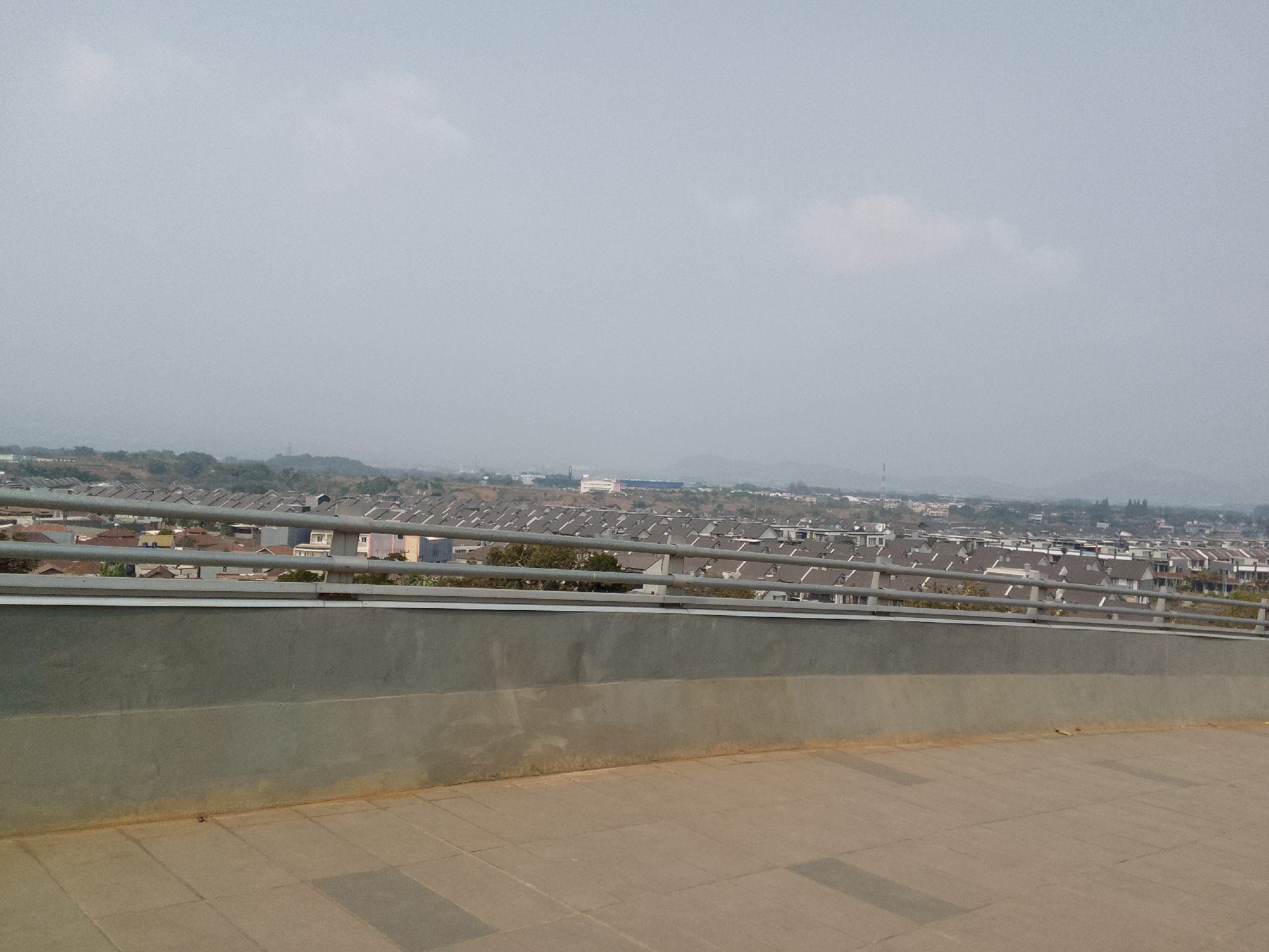 The image shows a view of the city of Bojonghaleuang, Indonesia, from a raised vantage point. In the foreground, a gray concrete wall with a silver railing runs horizontally across the frame. Beyond the wall, a dense residential area with many houses with gray roofs stretches into the distance. Some trees are visible interspersed among the houses. In the far background, a hazy landscape with hills and a hazy sky can be seen. The scene appears to be captured during the daytime. There are no people or active events depicted in the image.