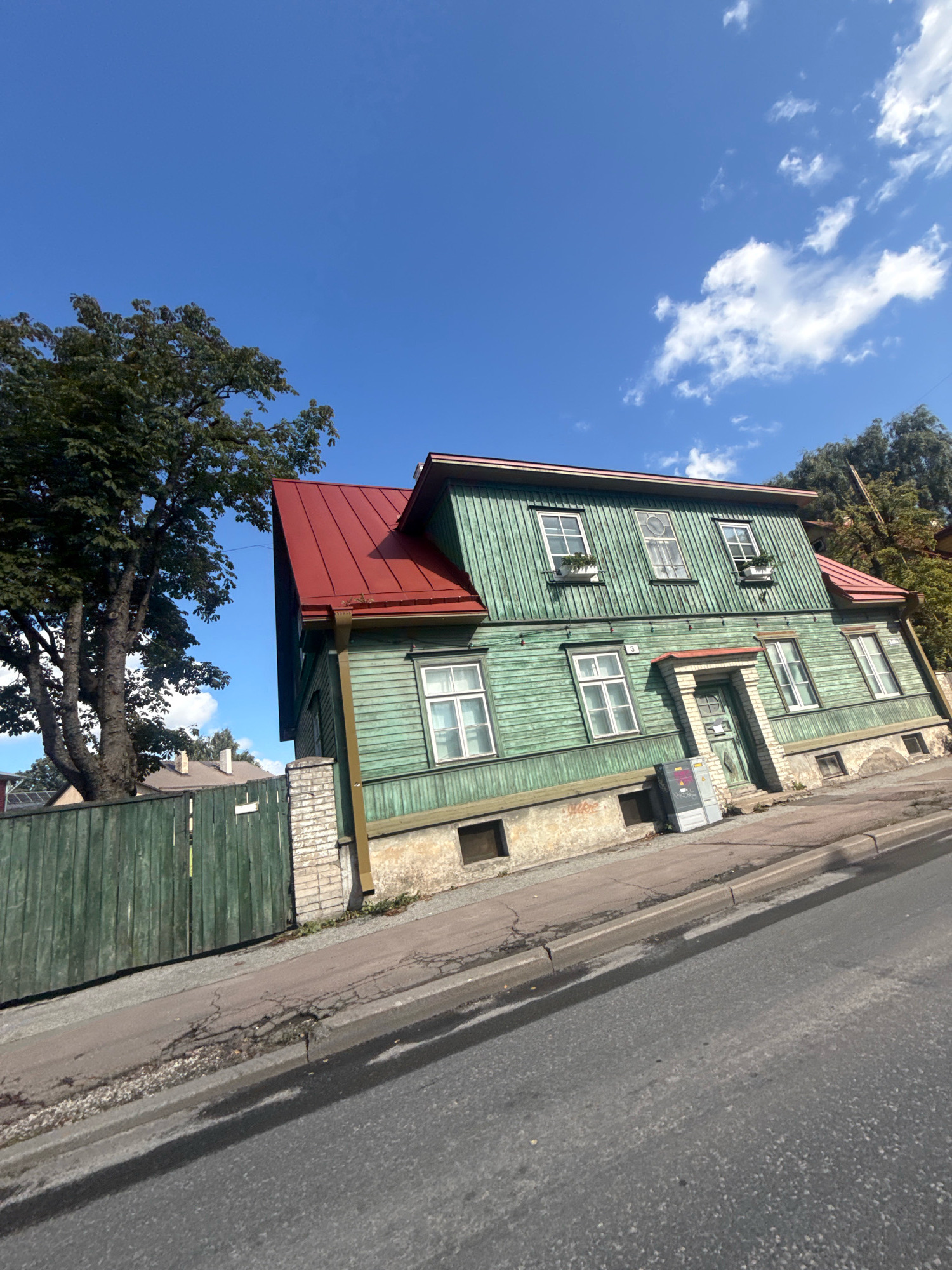 A bright, sunny day in Tallinn, Estonia features a two-story, light green wooden house with a red metal roof. The house is situated on a slight incline, with a grey asphalt road and sidewalk in the foreground. A tall, leafy tree with a thick trunk stands to the left of the house, behind a weathered green wooden fence. The house has multiple windows, some with small flower boxes, and a central entrance with a red-roofed awning. A grey utility box is visible next to the entrance. The sky is a vibrant blue with scattered white clouds.