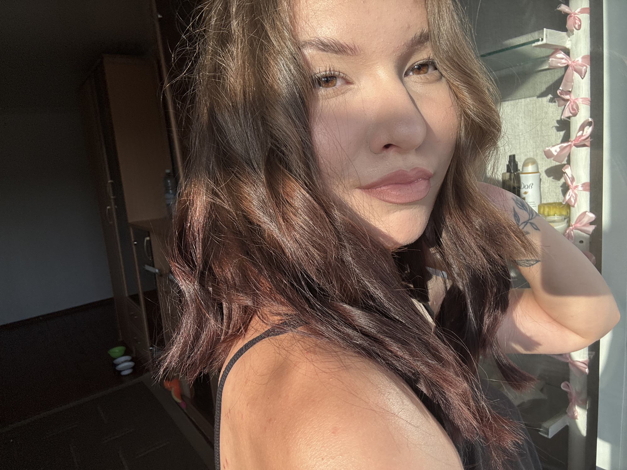 This is a medium shot, taken from a high angle, of a young woman with long, wavy brown hair. Sunlight casts a strong shadow across her face, illuminating her right cheek and eye. She is wearing a dark-colored top with a thin strap visible on her left shoulder. A tattoo is visible on her upper arm, and her skin appears smooth and slightly tanned.
In the background, to the left, is a dark room with a wooden cabinet or closet. The cabinet doors are slightly ajar, and a bottle of water and some stacked white and green objects are visible on the floor. To the right, a shelf unit is visible, adorned with small pink bows tied around its edge. Several cosmetic products, including a bottle with a black dispenser and a white aerosol can with "Dove" printed on it, are arranged on the shelf. The lighting suggests it is daytime, likely late afternoon given the warm sunlight. The setting appears to be indoors, possibly a bathroom or dressing area, in Inskoy, Russia. The woman's expression is neutral, with a slight gaze towards the camera.
