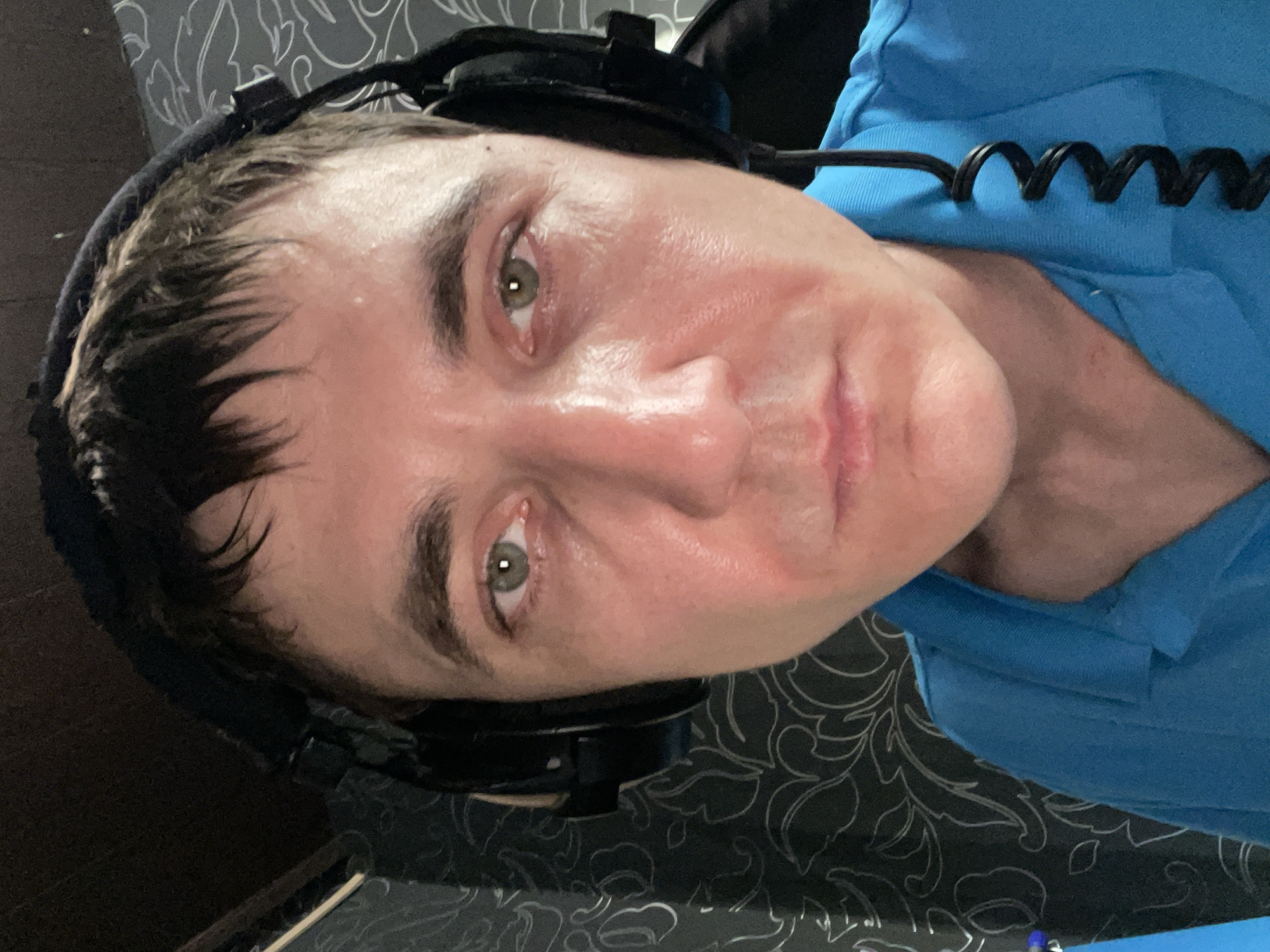 A man wearing black headphones and a blue collared shirt is shown in a close-up, forward-facing shot. His skin appears slightly shiny, possibly from sweat. The background consists of patterned wallpaper. The man is indoors. There is no discernible urban context or elements identifiable with Laishevo, Russia in the image.