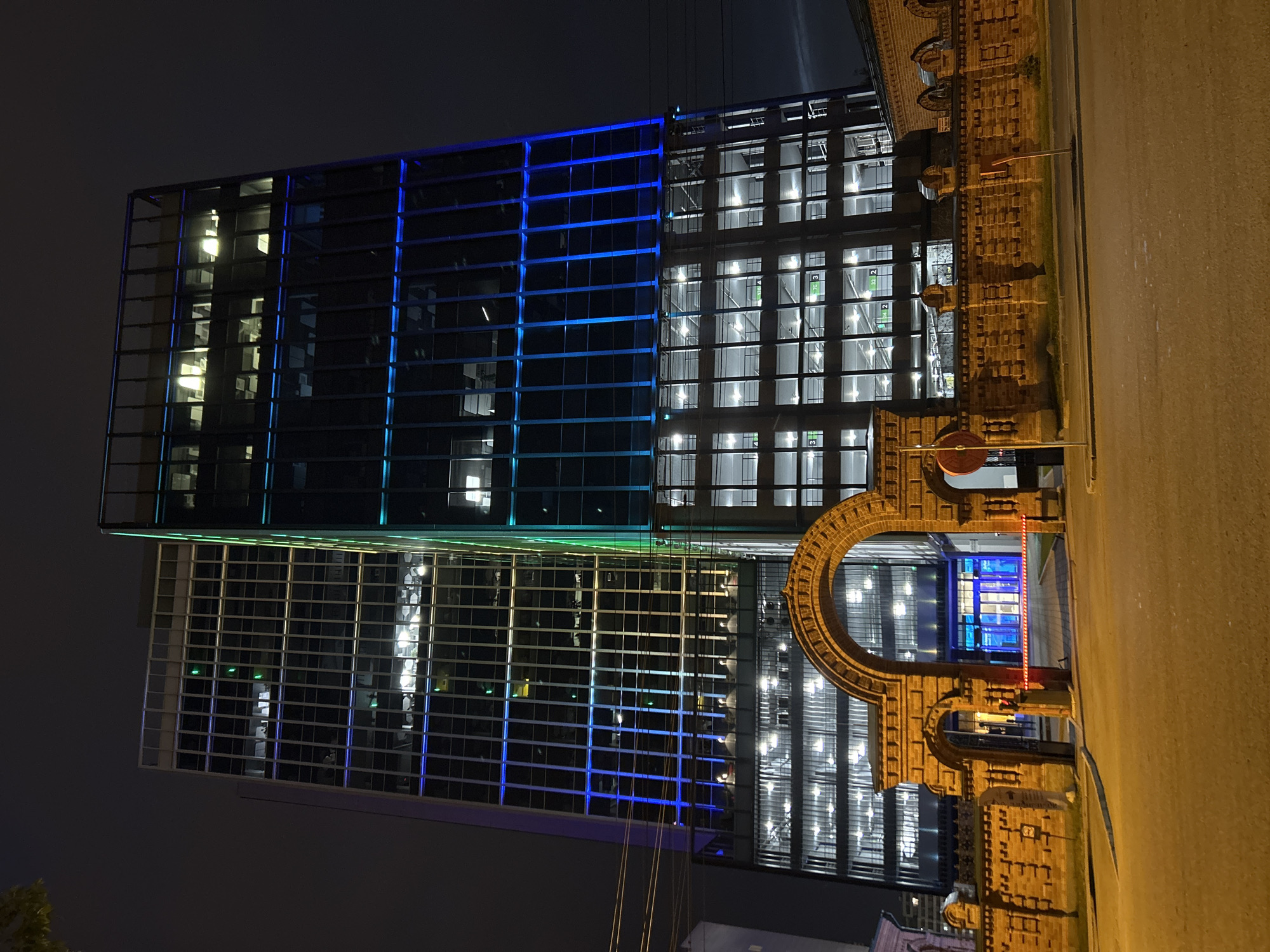 The image depicts a night scene in Yekaterinburg, Russia, showcasing two modern high-rise buildings with illuminated facades. The buildings are situated behind an old brick gate and wall structure, which frames the entrance to the property. The taller building on the right features blue and green accent lighting along its vertical panels, while the building on the left displays blue and yellow lighting. Both structures have numerous windows, many of which are lit from within, suggesting occupancy or active lighting on multiple floors. The foreground shows a paved road and sidewalk, with the brick gate and wall dominating the lower portion of the frame. The sky is dark and overcast. The scene is static and appears to be taken from a street-level perspective.
