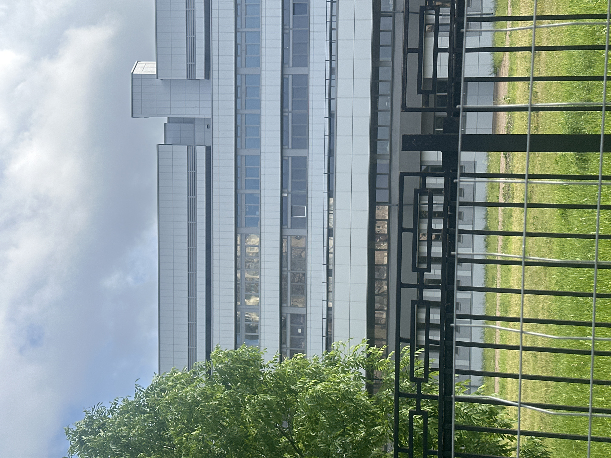 A medium shot captures a modern, multi-story building with a white facade and horizontal windows, partially obscured by a lush green tree on the left and a black metal fence in the foreground. The building appears to be a large institutional or commercial structure, with multiple levels of uniform windows reflecting the cloudy sky. The fence is composed of vertical bars and a decorative geometric pattern at the top. Behind the fence, a patch of green grass and a dirt path are visible, suggesting an outdoor space adjacent to the building. The sky is overcast with some patches of blue.