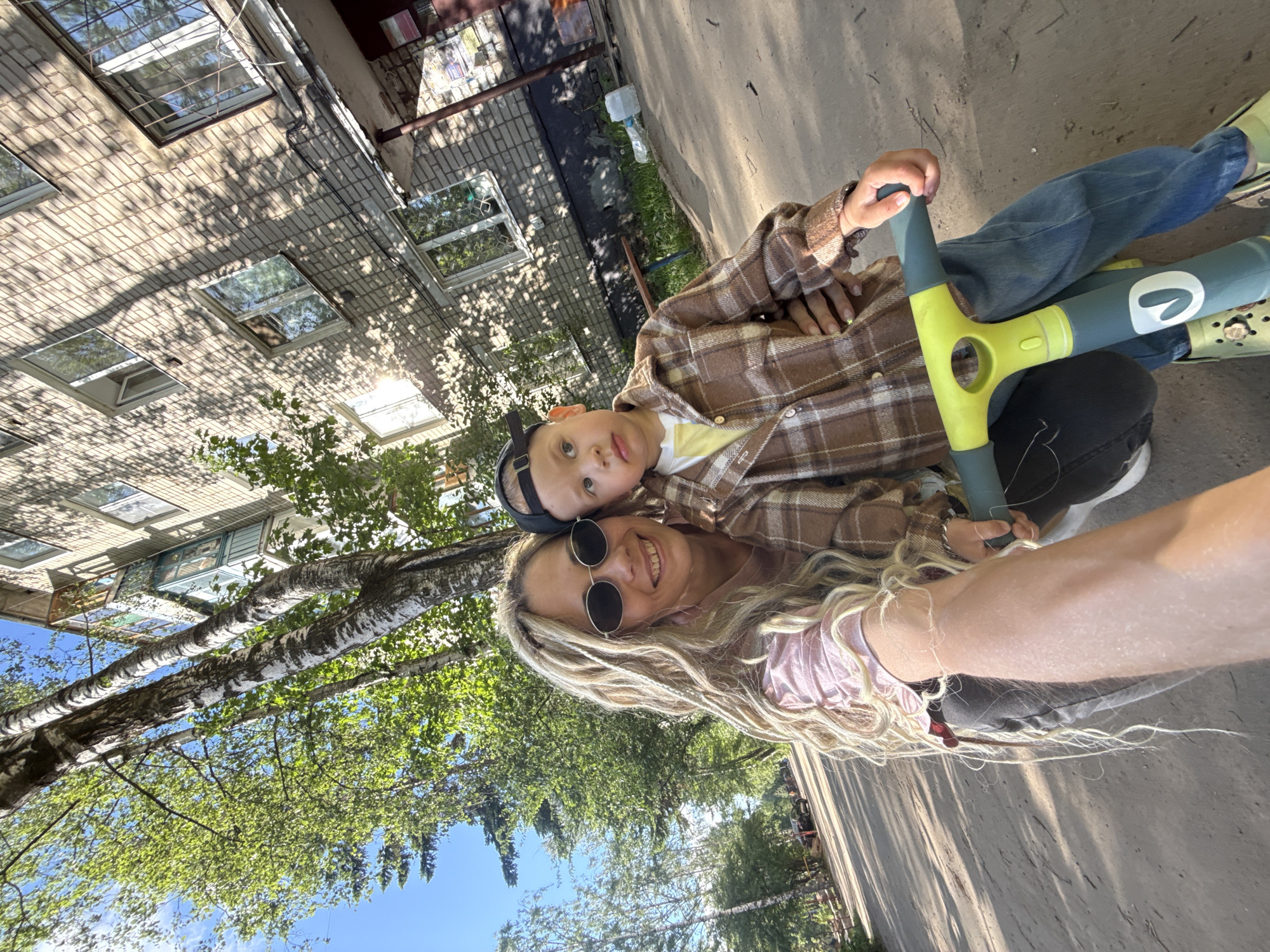 A selfie taken in Rusanovo, Russia, shows a woman with long blonde hair and round sunglasses holding a child on a scooter. The child is wearing a brown plaid shirt and a black baseball cap. The scooter is green and gray. They are standing on a paved surface near trees and a building with multiple windows. The sky is blue with some clouds visible through the trees. The photograph has a tilted perspective.