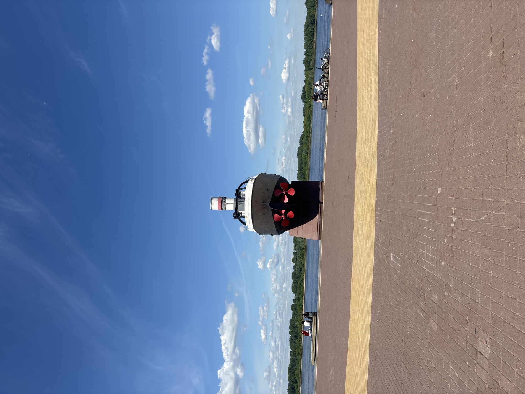 The image displays a bright, sunny day along a wide river, likely in Nizhny Novgorod, Russia. In the foreground, a spacious paved promenade or dock extends, composed of reddish-brown brick pavers transitioning into a lighter tan section closer to the water.
A prominent feature is a large, partial boat or ship structure positioned on the paved area, facing the river. This structure, appearing to be a monument or static display, is primarily white and black, with striking red propellers visible on its side and a mast-like element at its front.
Beyond the paved area lies a broad expanse of blue river water. The opposite bank is densely covered with a line of vibrant green trees. The sky overhead is a clear blue, adorned with scattered white cumulus clouds and some delicate, wispy cirrus formations, indicating pleasant weather conditions.
Several small figures are visible near the water's edge on the promenade. Some are seen next to bicycles, suggesting leisure activities such as cycling or enjoying the riverside view. Due to the distance, specific details of the individuals or their emotions are not discernible. The bright sunlight suggests it is likely midday or early afternoon. There is no visible text in the image. The scene conveys a sense of calm and outdoor enjoyment.