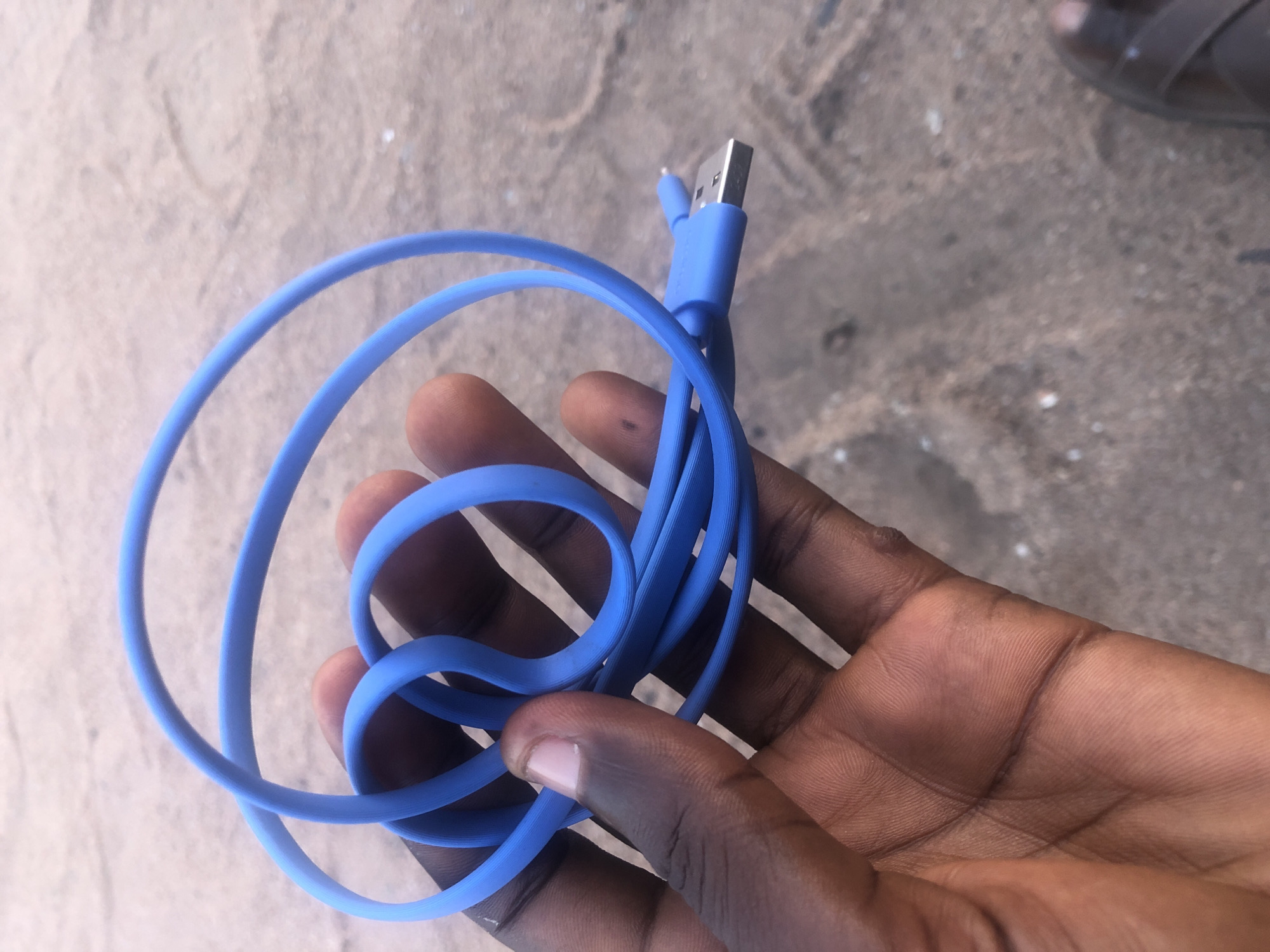 A person's hand, with dark skin and visible fingerprints, holds a coiled blue USB cable. The cable has a USB-A connector attached to one end. The background appears to be a sandy or dusty surface, possibly a road or ground in Jalingo, Nigeria, given the context provided. A brown sandal is visible in the lower right corner of the image. The lighting suggests it is daytime.