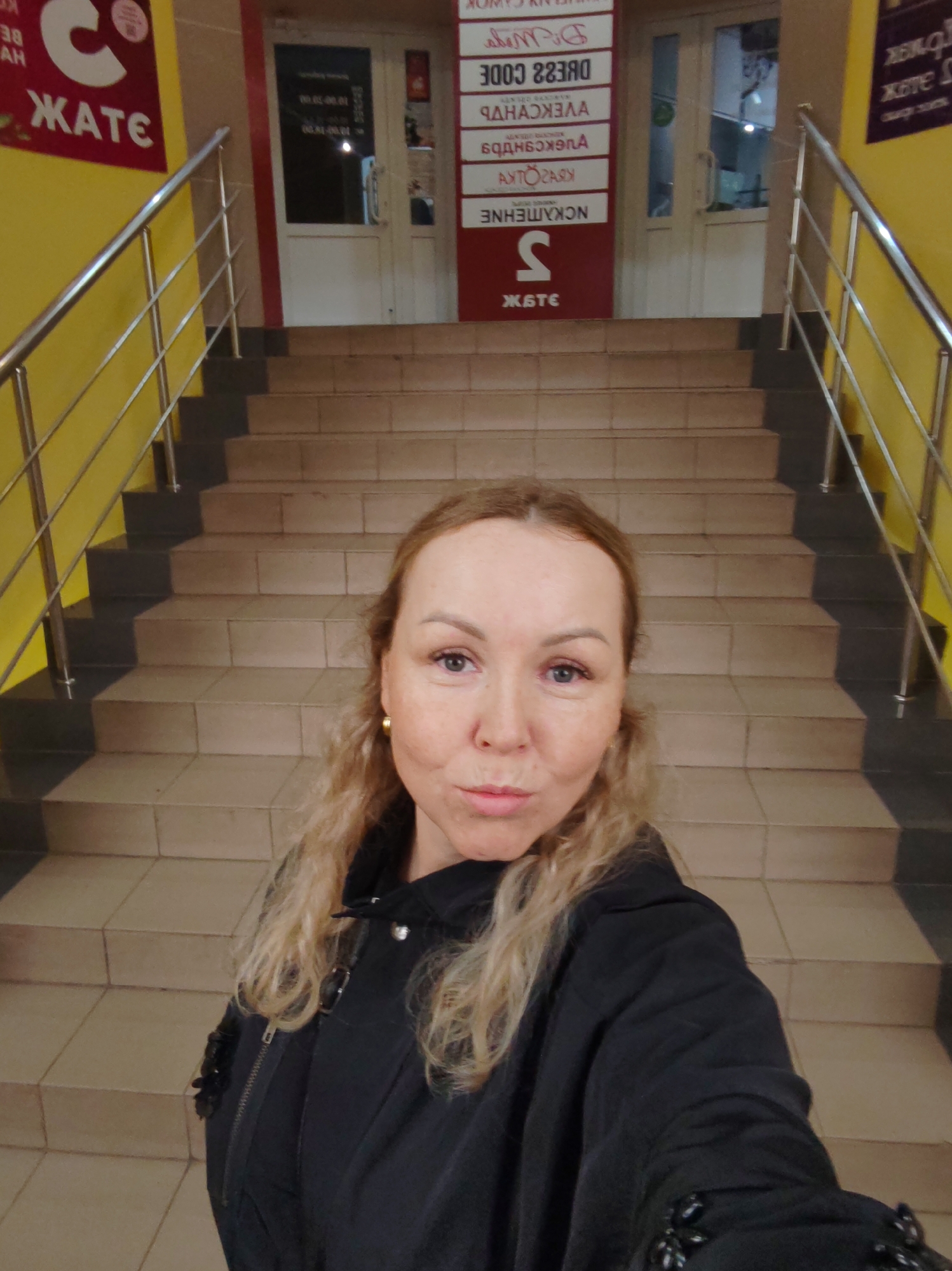 The image shows a woman taking a selfie on a staircase inside a building. The staircase has a yellow wall on one side with a metal railing. Above the staircase are signs in Russian, with text including "ЖАТЕ", "DRESS CODE", and "NCKAMEHNE". The location is Chaikovsky, Russia, based on the Russian text and other contextual clues. The woman is wearing a black jacket and has blonde hair. The environment is an indoor public space, possibly a shopping mall or commercial building.