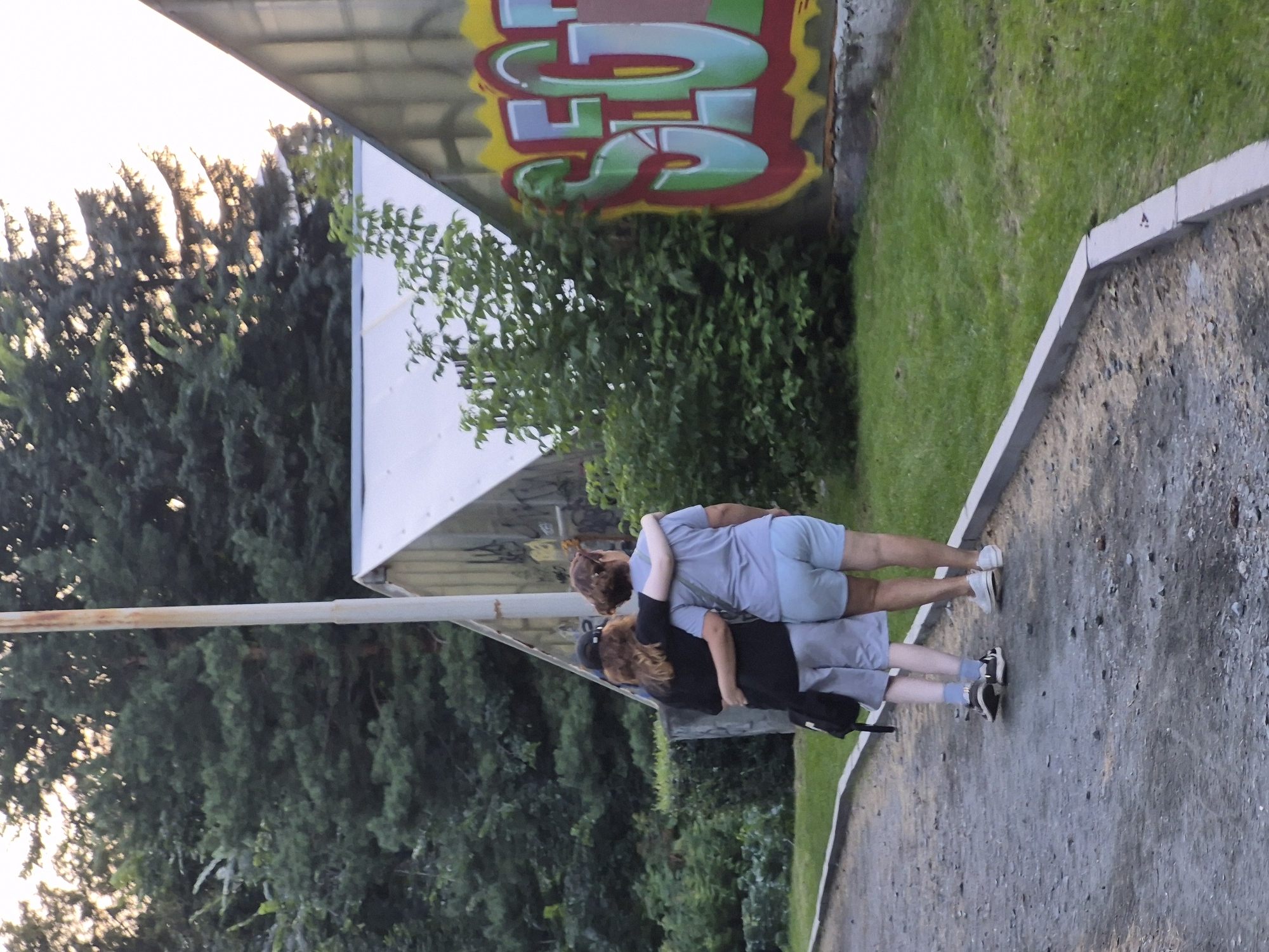 The image shows two people walking away from the camera on a gravel path. The person on the left is wearing a black shirt, grey shorts, and a black baseball cap. They have their arm around the person on the right, who is wearing a light blue t-shirt and light blue shorts. The path is lined with a concrete curb and grass. In the background, there are trees, a white structure with a pointed roof, and a building with graffiti on its side. The graffiti appears to be in red, yellow, green, and blue, with the letters "SEG" prominently visible. The lighting suggests it is late afternoon or early evening. There is no specific visual information in the image to ascertain the location as Yekaterinburg, Russia.