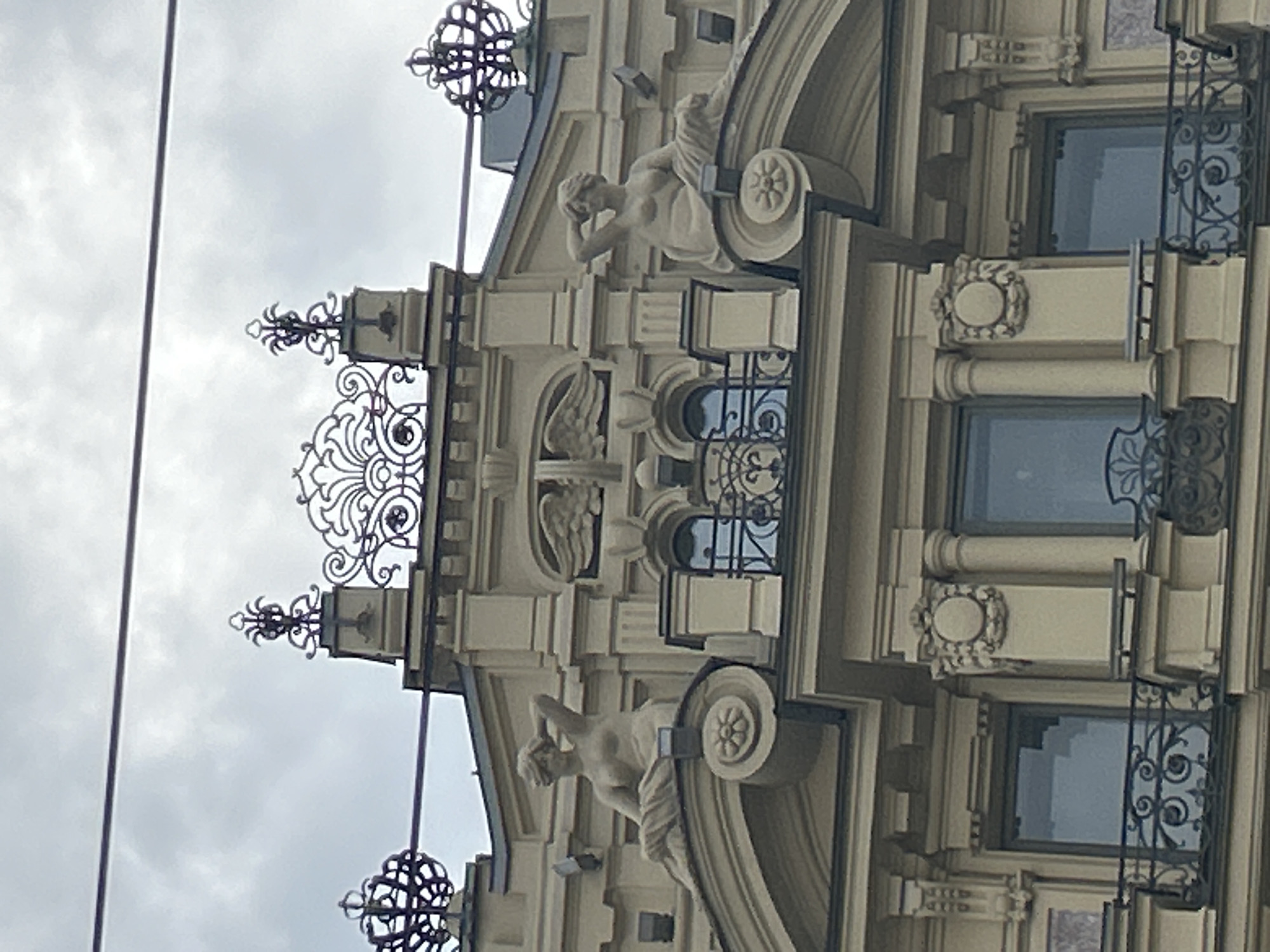 The image shows a close-up, low-angle view of the upper facade of an ornate, light-colored building, likely in an urban setting, with a cloudy sky in the background. Two sculpted female figures, nude from the waist up and draped in fabric below, are positioned on rounded architectural elements on either side of a central section. The figure on the left reclines with her left arm behind her head and her right hand resting on her hip, while the figure on the right leans back, supporting herself with her left arm behind her head and her right hand resting on her lap. Between the figures, a central architectural element features a decorative ironwork crest above two arched windows, each with a black metal railing in front. Above this, on the roofline, are more decorative ironwork elements, resembling crowns or elaborate finials, positioned atop small structures. Below the sculpted figures and windows, the building continues with more classical architectural detailing, including columns, cornices, and decorative reliefs, as well as balconies with intricate black metal railings. The overall impression is one of classical and baroque architectural styles.