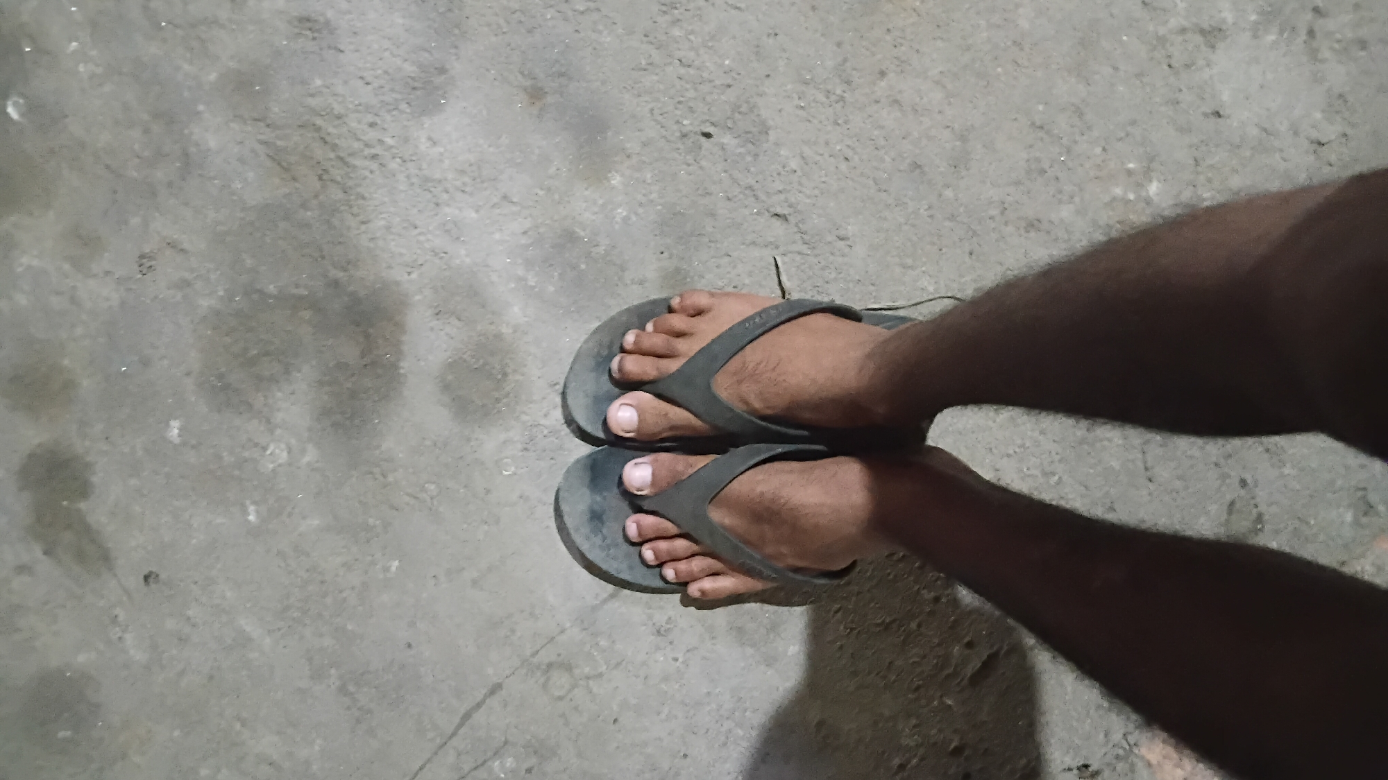 The image shows a pair of feet wearing black flip-flops on a concrete surface. The feet are tanned and hairy, and the toenails are visible. The concrete surface appears to be outdoors, possibly in a public space in Prabumulih, Indonesia, given the context of the prompt. There are some smudges and possibly water stains on the concrete. The lighting suggests it is nighttime or a dimly lit area. The image is taken from a top-down perspective.