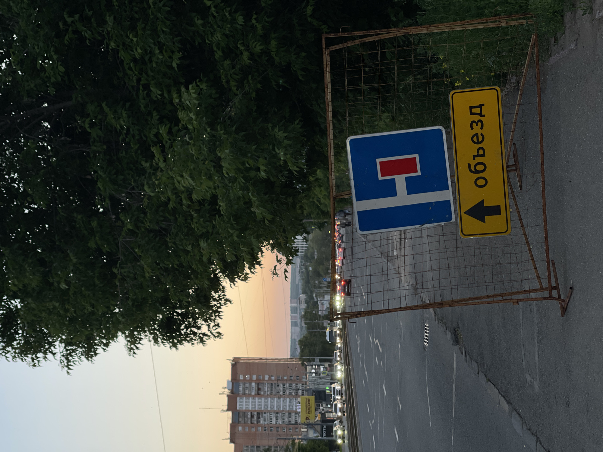 A road scene is depicted at dusk. In the foreground, a temporary metal fence with wire mesh is erected, holding two road signs. The upper sign is blue with a white horizontal bar and a red rectangle. The lower sign is yellow with black text that reads "объезд" and a black arrow pointing upwards. The fence and signs are positioned on a sidewalk next to a multi-lane road with traffic. Cars are visible on the road, with headlights on, suggesting evening traffic. Across the road, there are buildings, streetlights, and trees. The sky in the background shows a soft gradient of orange and blue, characteristic of sunset. A large, leafy green tree dominates the right side of the frame, partially obscuring the background.