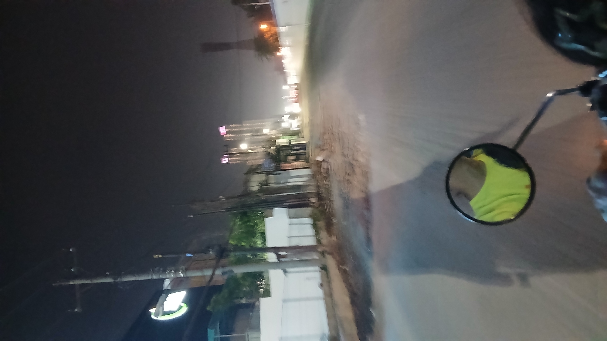 The image shows a nighttime view of a street in the City of Medan, Indonesia, as seen from the perspective of someone riding a motorcycle. In the foreground, a round rearview mirror attached to the motorcycle reflects the rider's face and a bright yellow t-shirt. The road ahead is paved and appears somewhat uneven. To the left of the road, a white wall runs along the side of a property, with a utility pole situated in front of it. Further down the street, several tall buildings with illuminated windows and pink accents rise into the night sky, suggesting a developed urban area. Streetlights and the lights from the buildings illuminate the scene. The overall impression is of a journey through a moderately lit urban environment at night.