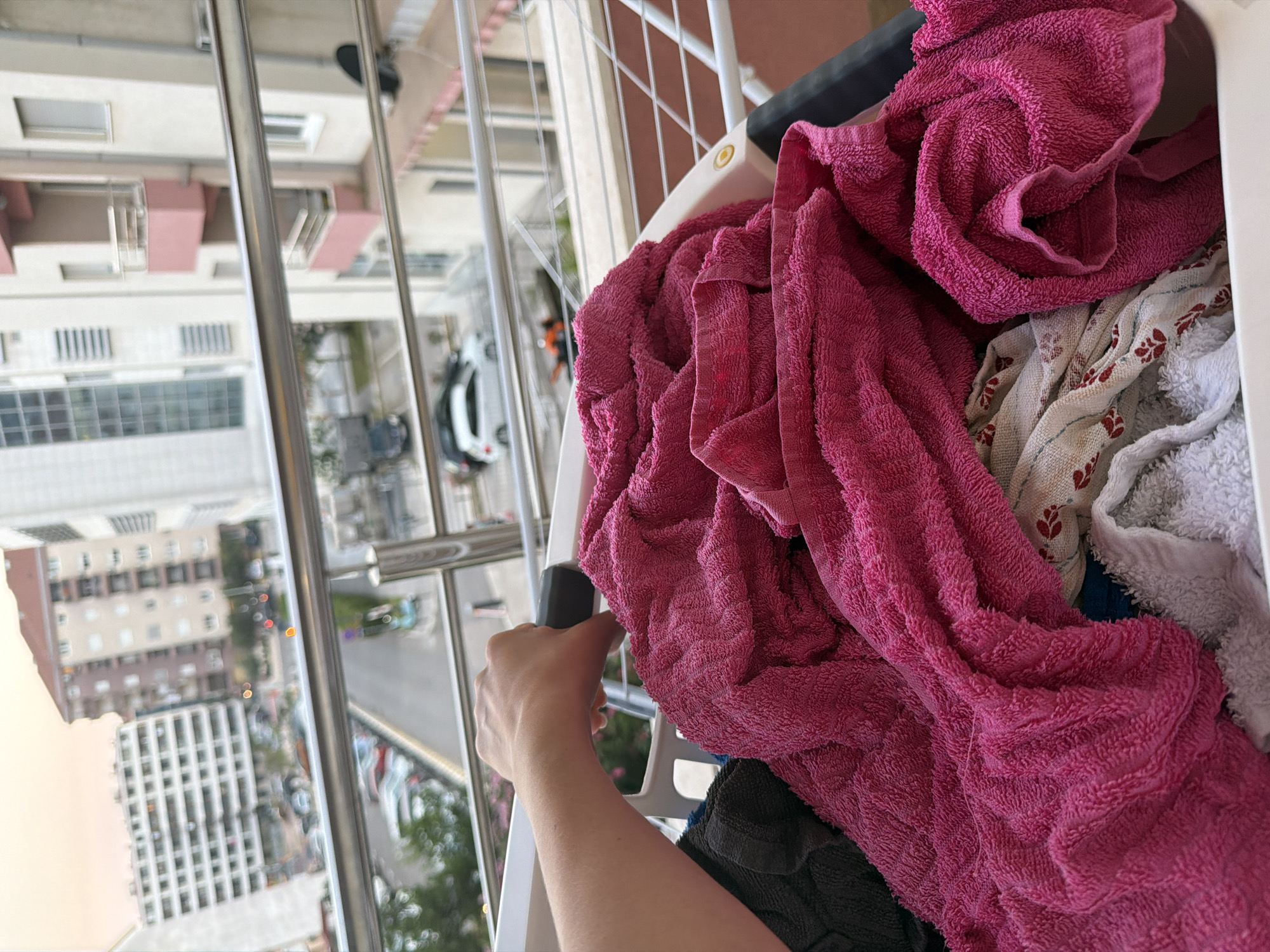 A close-up shot focuses on a person's hand gripping a clothes rack filled with laundry. The laundry includes several pink towels and some patterned fabrics. In the background, a balcony railing overlooks a street in Budva, Montenegro, with buildings, cars, and a pale sky.