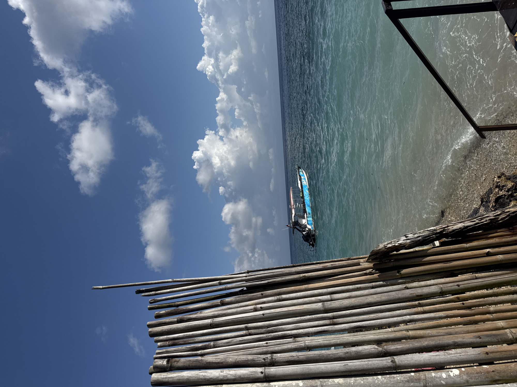 The image captures a scene from Nusa Penida, Indonesia. The foreground features a bamboo fence or barrier constructed of closely packed bamboo poles running horizontally across the lower portion of the frame. The fence appears to be aged and weathered.
Beyond the fence, the view opens up to the ocean. The water is a clear, light turquoise color closer to the shore, fading into darker blues in the distance. A windsurfing board with a blue sail is visible in the water, seemingly unoccupied.
On the right side of the frame, a portion of a dark, possibly metal, structure intersects the scene at an angle. The shoreline consists of small pebbles and rocks.
The sky above features a clear blue sky with scattered white cumulus clouds. The composition suggests a coastal or beach location, with the bamboo fence serving as a boundary or marker. The angle of the shot is such that the scene appears rotated counterclockwise.