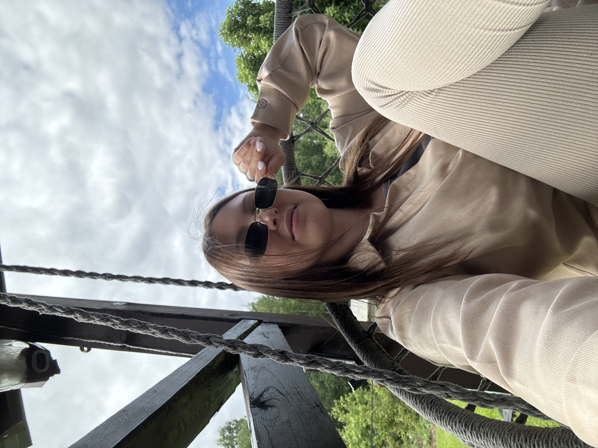 A young woman with long brown hair, wearing sunglasses, a beige collared shirt, and cream-colored corduroy pants, is seated in a woven chair hanging from ropes. She is looking towards the camera with her right hand touching her sunglasses. The background shows green trees and a cloudy sky. The scene appears to be outdoors, possibly on a porch or balcony. This image was taken in Ponasirevo, Russia.