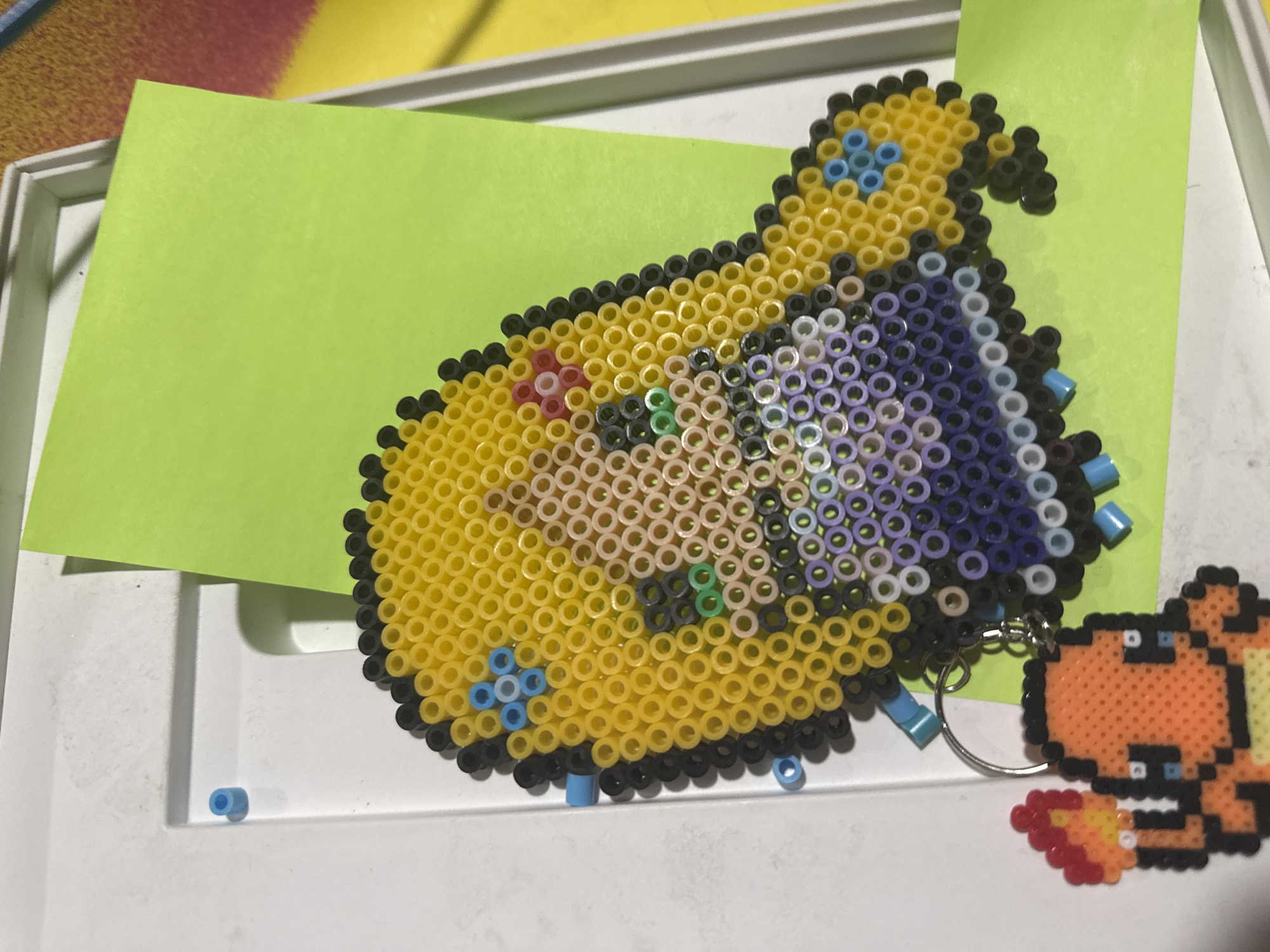 The image displays two Perler bead creations placed on a bright lime-green piece of paper, which rests inside a white plastic tray or frame.
The main subject is a large, colorful Perler bead art piece. It is shaped like a stylized object or character, predominantly yellow with a black outline. Internal details are rendered with light purple, beige, light pink, light green, and light blue beads, creating intricate patterns within the yellow form. Towards the upper right, the piece transitions into a darker purple and black section, suggesting a contrast or different material.
To the bottom right of the larger piece, a smaller Perler bead creation is visible, depicting Charmander, a Pokémon character. This Charmander is primarily orange with black outlines, white eyes, blue pupils, and a flame at its tail made of red, orange, and yellow beads. It is attached to a metallic keyring, indicating it serves as a keychain.
Several loose blue Perler beads are scattered on the white tray surface near the creations, suggesting they might have recently been crafted or are part of an ongoing project. The background includes glimpses of other colored surfaces, possibly a table or mat, in hues of yellow and red. No people or visible text are present in the image. The time of day and weather cannot be determined. The location context for this media file is Puebla, Mexico.