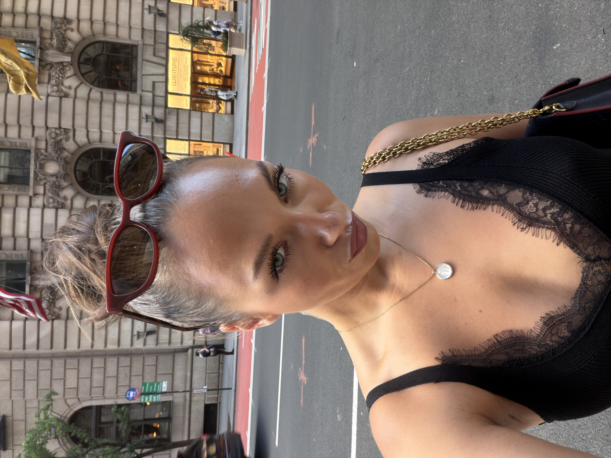 A woman takes a selfie on a city street, likely in New York City, USA. She has her hair pulled up and red sunglasses perched on her head. She is wearing a black camisole with lace detailing and a gold necklace. Behind her, a building facade with ornate architectural features is visible, along with windows and awnings. A street with red markings and a crosswalk is in the foreground, with a few pedestrians and a car in the background. The overall setting suggests an urban environment with a mix of classic architecture and street life.