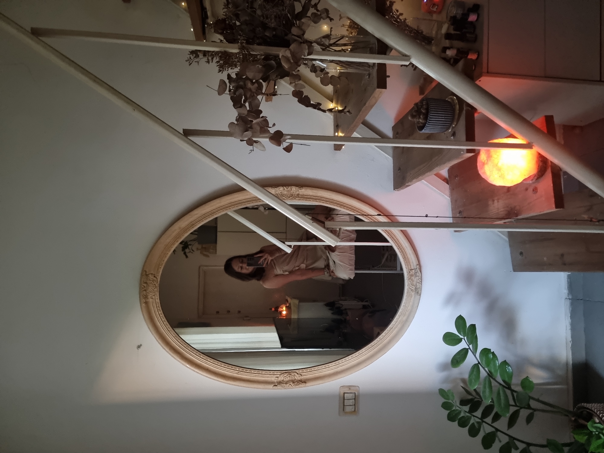 A woman is reflected in an oval mirror hanging on a white wall. She is holding a smartphone, presumably taking a photo of herself in the mirror. The mirror is positioned near a staircase with wooden steps. On the steps, there is a collection of items, including a lamp emitting a warm orange light, a plant, and a decorative pot. Near the bottom of the mirror, there is a light switch on the wall. A plant stands on the floor below the mirror. The image may have been taken indoors in a location in Tel-Aviv, Israel, but there is no definitive information to confirm this.