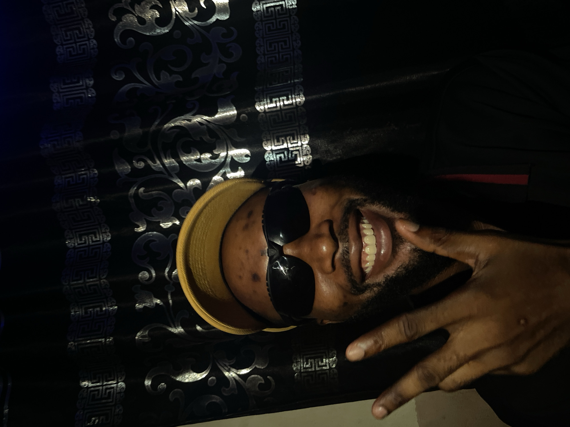 A person with dark skin, wearing a yellow cap and sunglasses, smiles and makes a hand gesture against a dark, patterned background. The person appears to have a beard. The background has silver or metallic designs. The scene is dimly lit. There is no verifiable information to locate the city of Lokoja, Nigeria.