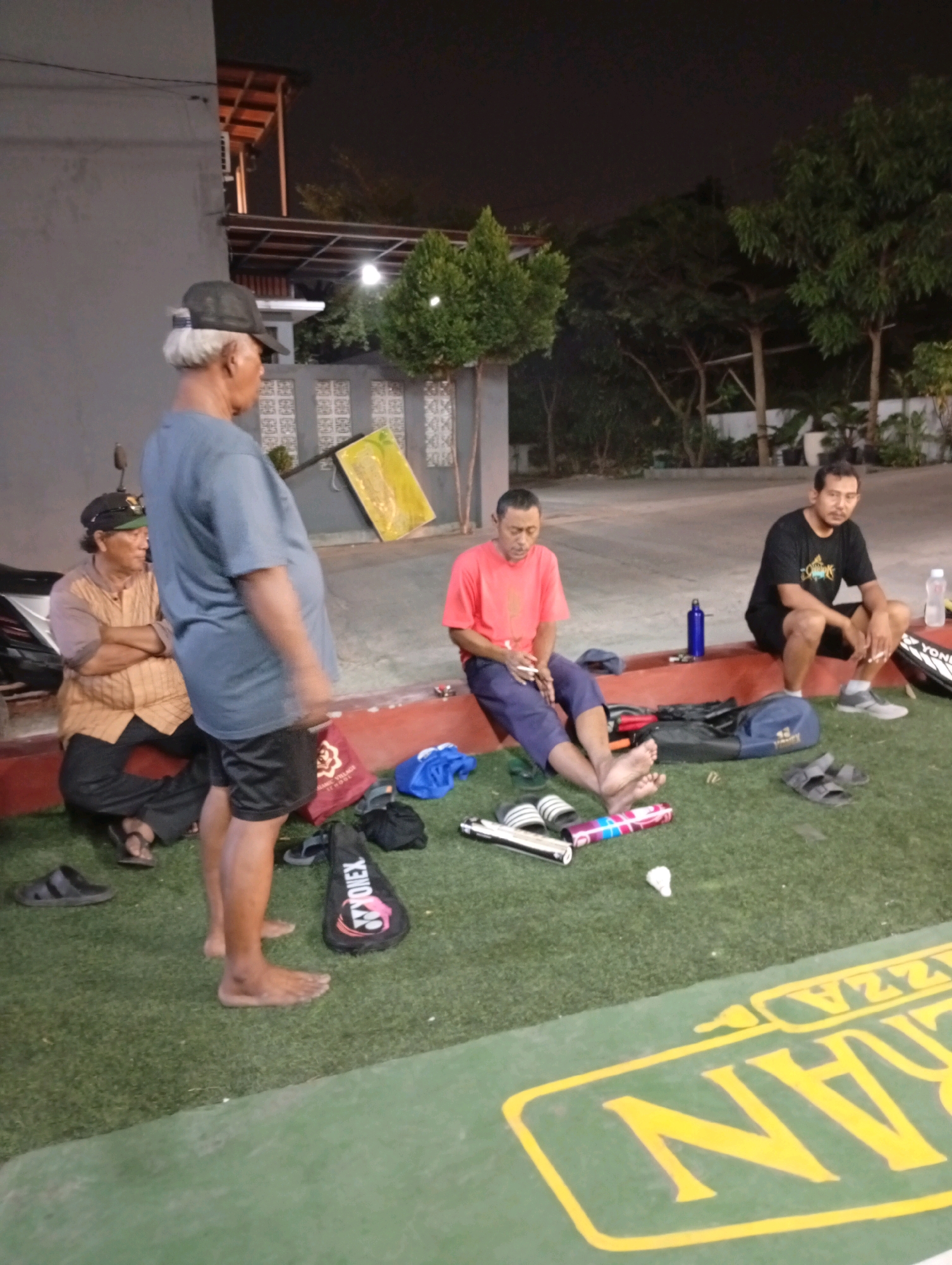 In an outdoor recreational area in Caringin, Indonesia, three men are gathered in the evening. One man, wearing a blue t-shirt and black shorts, stands with his back to the camera. To his left, another man in a brown shirt and black pants sits with his arms crossed, wearing a dark baseball cap. Further to the right, a man in a red t-shirt and blue pants sits on a ledge, holding a cigarette. Adjacent to him, a man in a black t-shirt and shorts also sits, smoking. The ground is covered in artificial turf, and various items are scattered around, including badminton rackets, a blue duffel bag, a red bag, footwear, and water bottles. The scene is lit by artificial lights, casting long shadows. A building with a tiled roof and trees are visible in the background, suggesting the location is likely a park or sports facility.