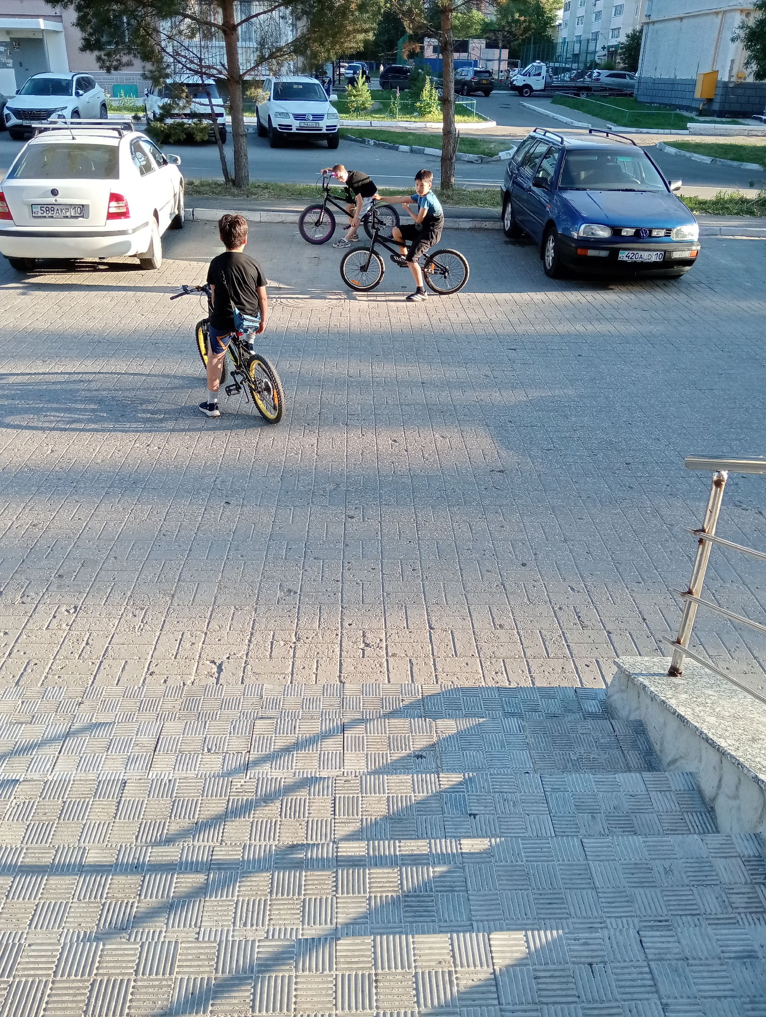 In an outdoor urban setting, three boys are present. One boy, facing away from the camera and wearing a black t-shirt and shorts, stands with a bicycle. Another boy, also facing away, is on a bicycle, wearing a black t-shirt and blue shorts. A third boy, to the right, is leaning over his bicycle, wearing a black t-shirt and dark shorts. They are on a paved area, with cars parked in the background. The environment appears to be a parking lot or street next to a building.