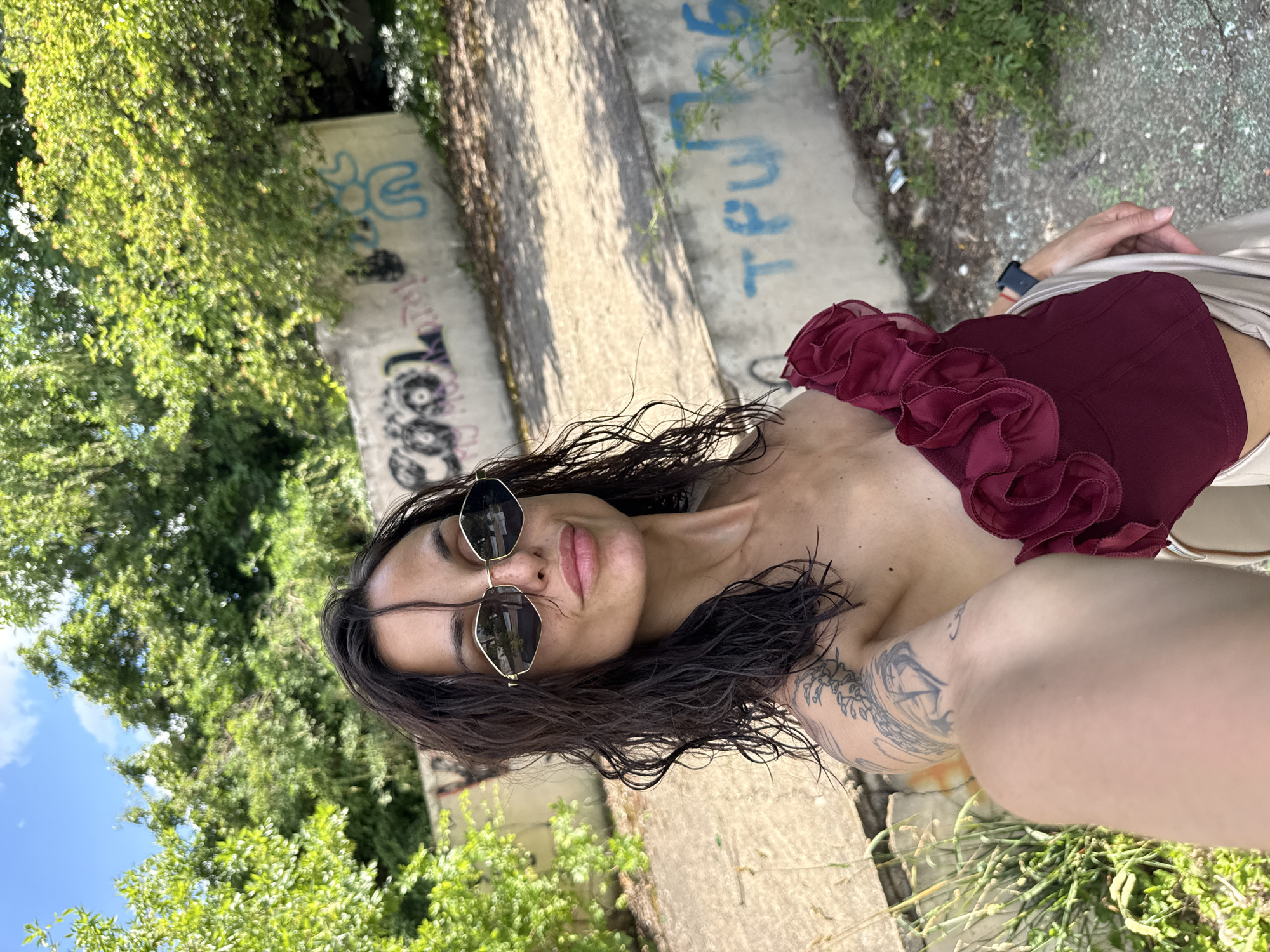 A woman with dark, wet hair, wearing sunglasses and a burgundy top, takes a selfie outdoors. The background consists of a concrete wall with graffiti and lush green trees under a blue sky with white clouds. The environment appears to be an urban setting, possibly abandoned or under renovation, judging by the graffiti and debris on the ground. The woman's pose suggests confidence and a casual demeanor. The overall lighting indicates a bright, sunny day. The location is Pleven, Bulgaria.