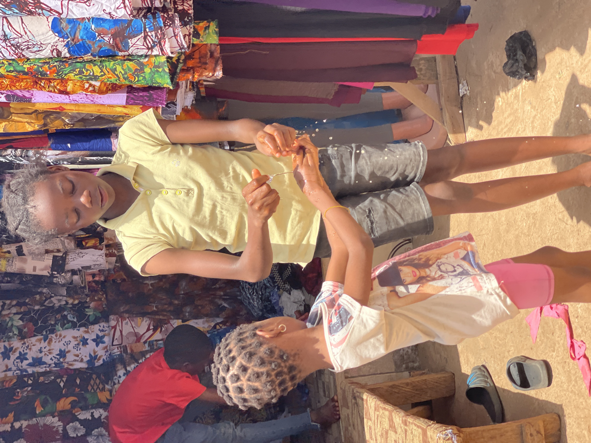 Two young girls are standing next to a fabric stall in Samaru, Nigeria. One girl is wearing a light yellow shirt and dark gray shorts. She is holding a thin, silver object and some droplets of water are falling from it. The second girl is wearing a white t-shirt with a graphic print and pink shorts. She is also looking at the object in the other girl's hands. Behind them is a stall filled with stacks of colorful fabrics. A person wearing a red shirt is behind the stall. The ground is dusty and there are shoes and a black plastic bag nearby.