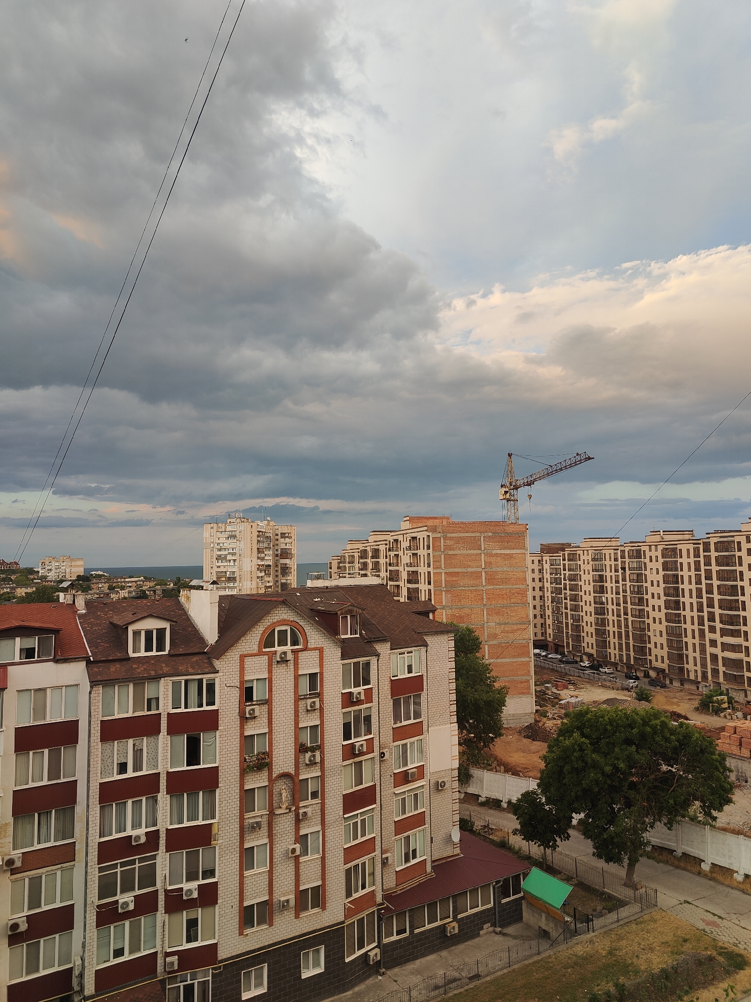 The image shows an aerial view of a residential area in Chornomorsk, Ukraine, under a cloudy sky. In the foreground, there are multi-story buildings with pitched roofs. One building features a red-brown roof and white and red brick facade, adorned with decorative elements and numerous air conditioning units. Adjacent to it is a lighter colored building with a similar roofline and facade. Further in the background, several large, modern apartment complexes are visible, characterized by their light-colored facades and numerous balconies. A construction crane stands tall next to an unfinished brick building, indicating ongoing development. The sea is visible on the horizon in the distance. A few trees are scattered throughout the scene, and a street with parked cars runs alongside one of the apartment complexes. Overhead wires stretch across the sky, and a tiny bird is visible in the upper left corner. The overall impression is of an urban environment undergoing construction and modernization, with a backdrop of a coastal city.