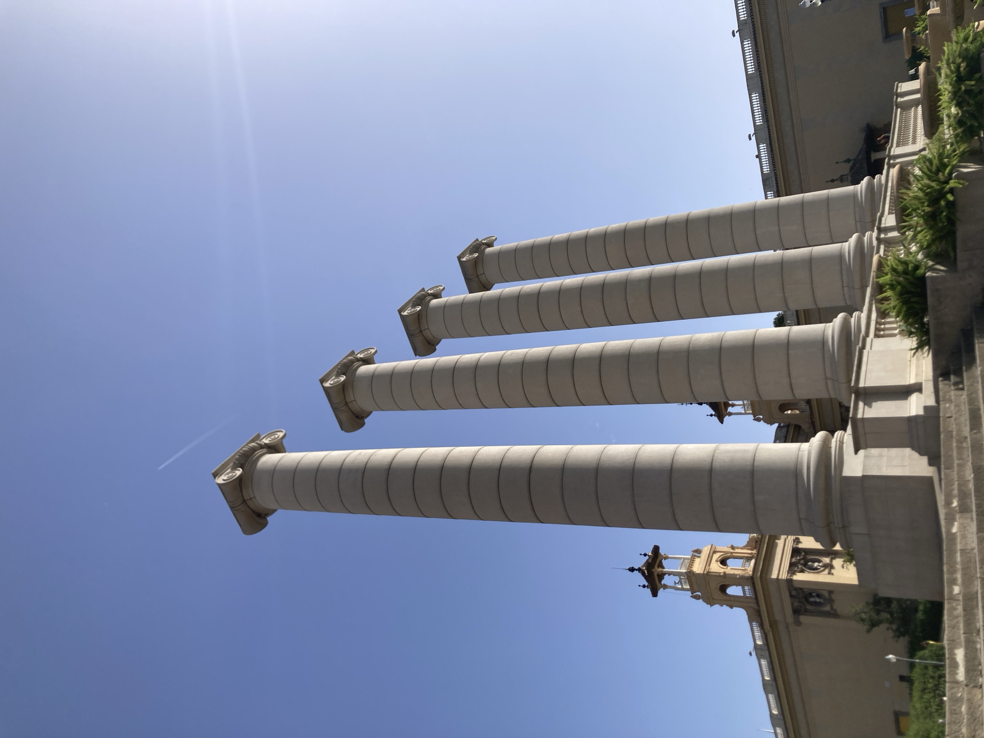 The image depicts the Four Columns of Carles Buïgas, a monument located on the Avinguda de la Reina Maria Cristina in Barcelona, Spain. Four monumental classical columns stand tall against a clear blue sky. The columns are cylindrical, fluted, and topped with Ionic capitals. In the background, a beige building with decorative architectural elements, including a small tower with a roof, is visible. Green foliage and steps are present in the foreground. A contrail from an airplane is visible in the sky. The sun is shining brightly, causing a lens flare on the right side of the image. No people are clearly visible in the image.
