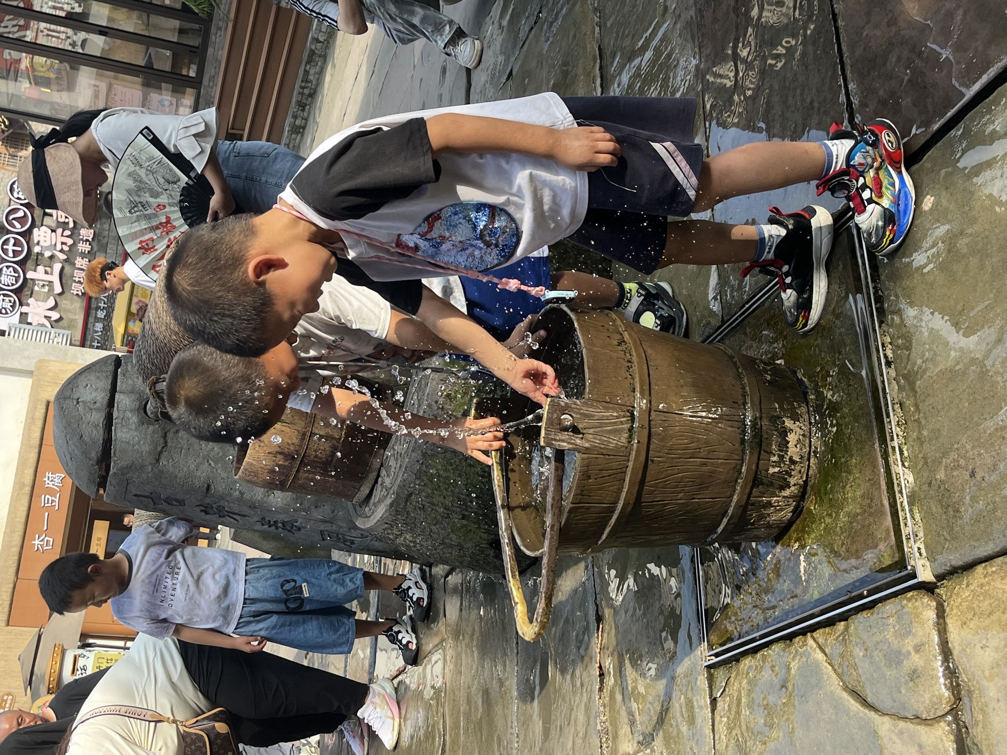 In Yuzhong District, China, a group of children is gathered around an outdoor fountain. The fountain features a large wooden bucket from which water is splashing. Two young boys are actively playing with the water, one using his hands to direct the flow, and the other reaching into the bucket. A third boy, wearing a t-shirt with a graphic design and dark shorts, stands to the right, his hands near the bucket. Another boy in the background, wearing a t-shirt and blue shorts, also appears to be interacting with the fountain. A woman with a fan stands behind the boys. The scene takes place on a wet, paved area with a metal grate around the fountain's base. The surrounding environment suggests a public space with shops and signage in the background, indicating an urban setting. The lighting suggests it is daytime.