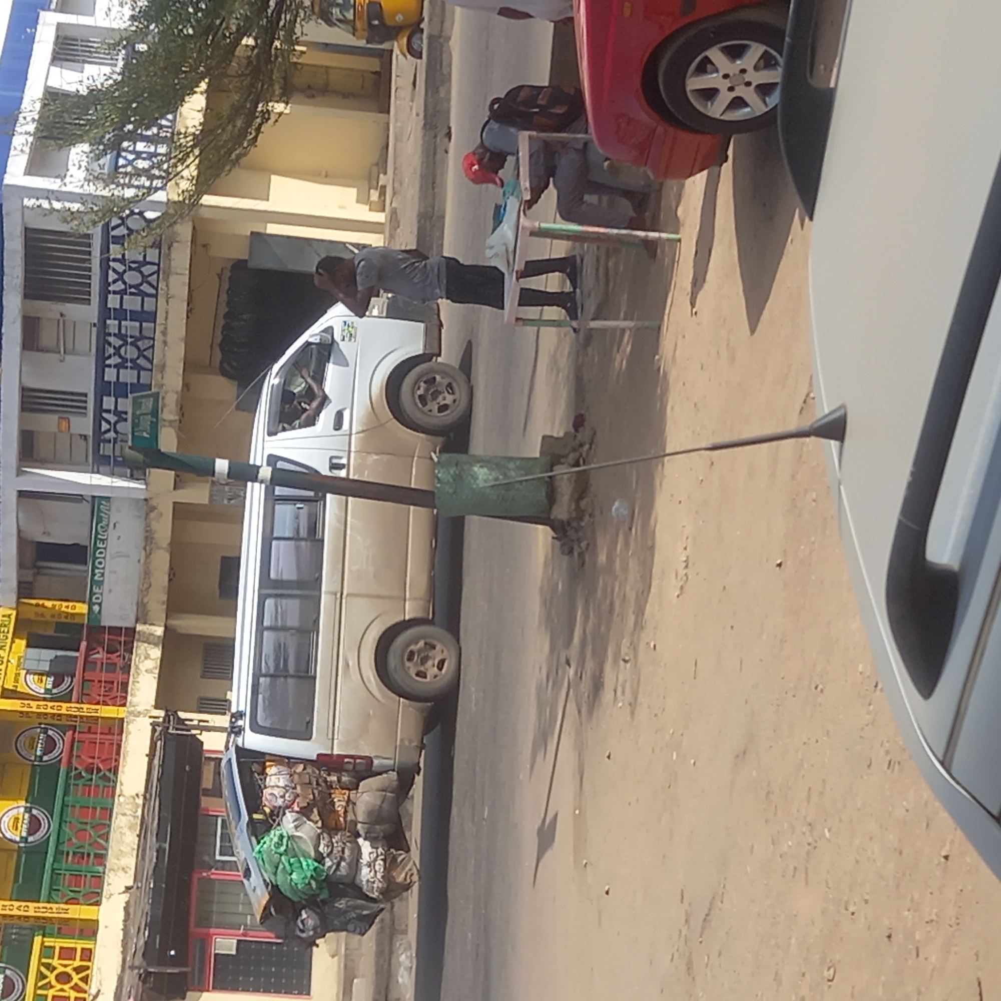 The scene appears to be located on a street in the city of Dadi Riba, Nigeria. A white van parked on the side of the road is heavily laden with goods, visible in the back. A utility pole with a green base stands near the van. Two men are present; one, wearing a grey shirt and dark pants, stands near a small table. The other man sits at the table, wearing a red hat and a grey shirt. A red car and a grey car are parked to the side. Various buildings line the street, including a structure with yellow, green, and red signage and another with a blue and white facade. The road is paved and dusty.