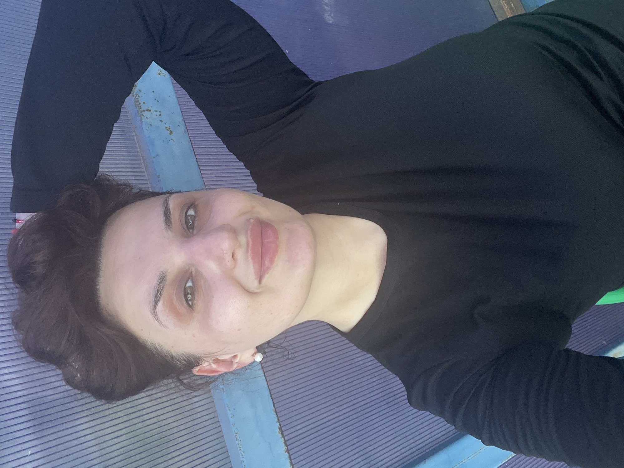 A medium shot captures a woman from the chest up, posing for a selfie against a backdrop of blue and purple corrugated plastic. Her dark hair is styled up, with some strands falling loose around her face. She wears a simple black long-sleeved shirt and a small white pearl earring in her visible ear. The woman's right arm is raised, with her hand resting on the back of her head, creating a relaxed pose. She looks directly at the camera with a gentle smile, her makeup subtle with a touch of blush and lip gloss. The lighting is natural, casting soft shadows and highlighting the texture of the corrugated background. The overall impression is one of casual comfort and self-assuredness.