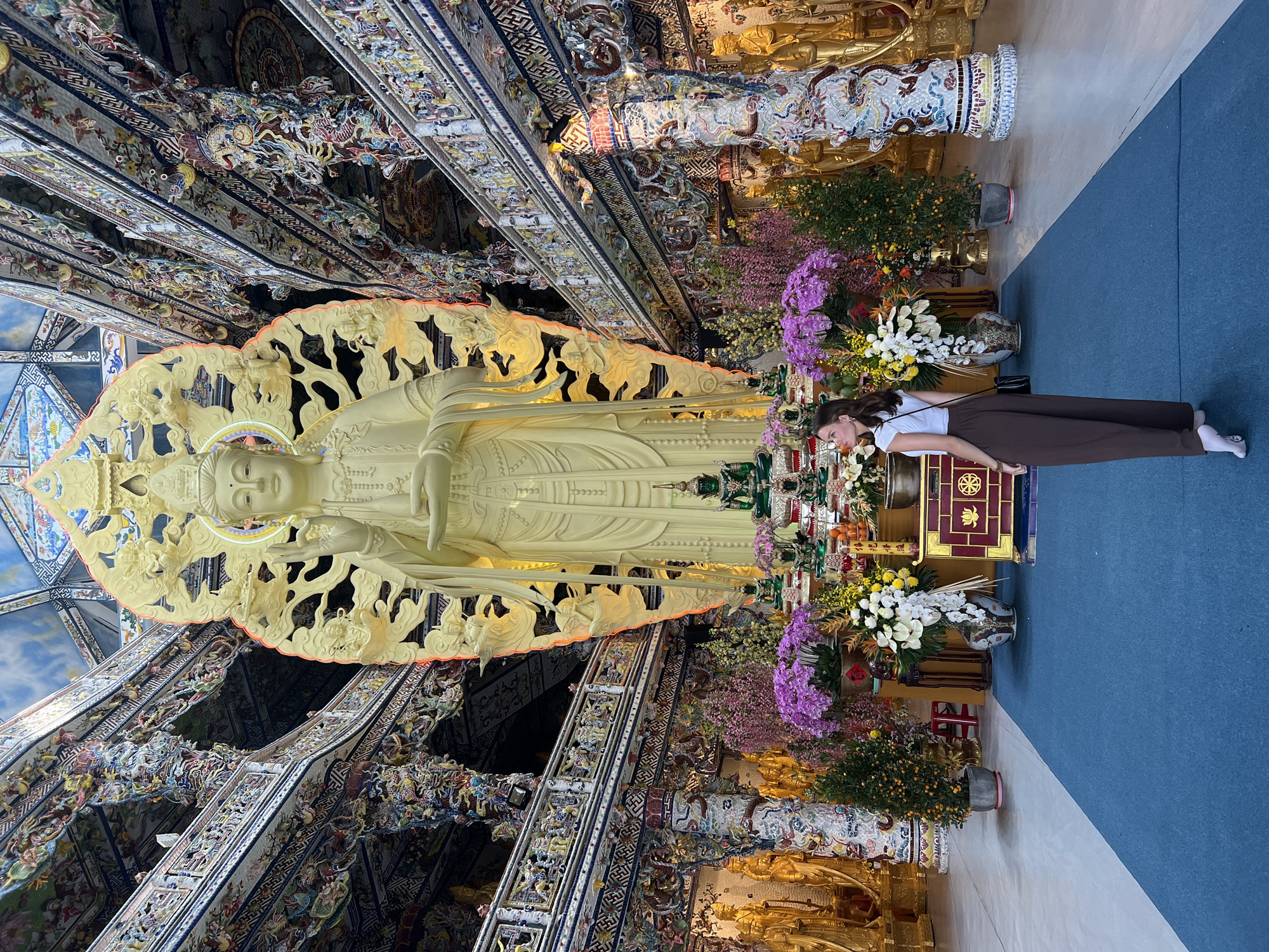 The image shows a tall, cream-colored Buddha statue in a temple in Xuân Thành, Vietnam. The statue is surrounded by a golden halo and intricate carvings on the ceiling and walls. A woman stands in front of the altar, near the statue, with her arms outstretched. There are vases of flowers arranged around the altar, and a blue carpet covers the floor in front of the altar.