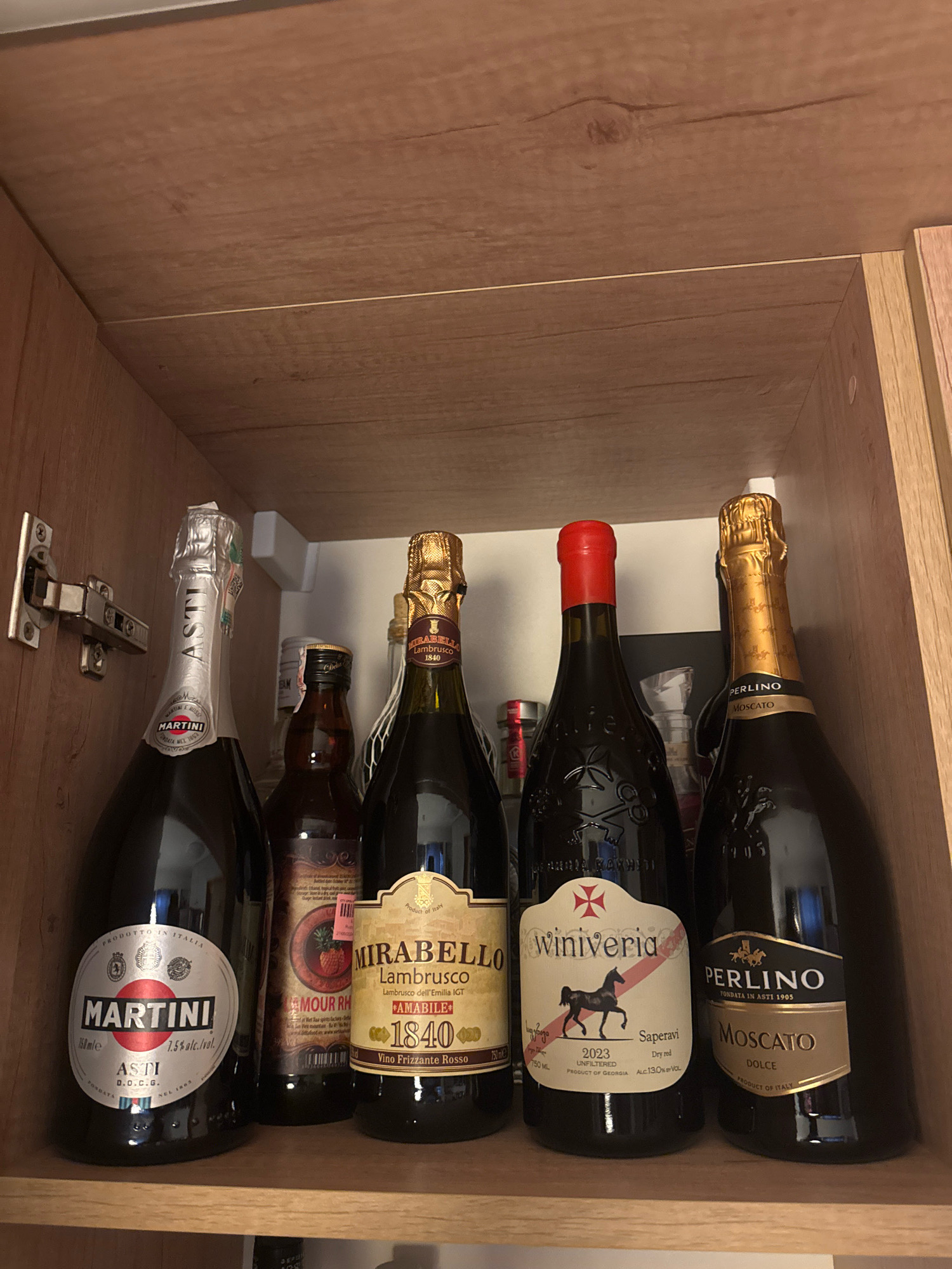 A wooden cabinet is open, revealing several bottles of alcohol on a shelf. The bottles include Martini Asti, a bottle with a label featuring a pineapple, Mirabello Lambrusco, Winiveria Saperavi, and Perlino Moscato. The cabinet appears to be in a home environment. The image is a still photograph and does not depict any specific location in Almaty, Kazakhstan.
