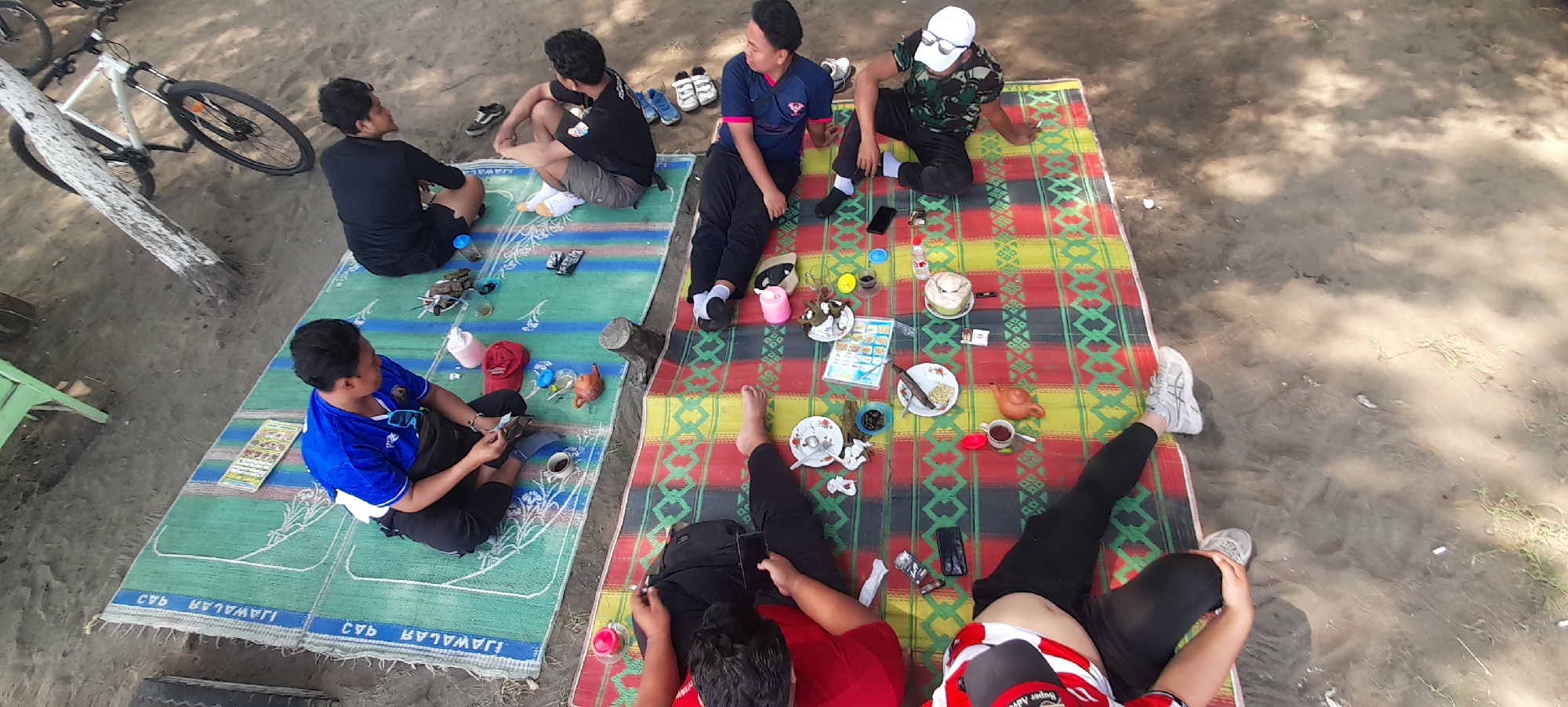 The image captures a group of five young men gathered on a beach in Nyamplung Sari, Indonesia. They are seated on two different mats spread on the sand, engaged in a casual picnic. The setting appears to be outdoors, likely during the daytime, with sandy terrain and some vegetation visible. The lighting suggests it could be late morning or early afternoon.
One mat is predominantly green with white patterns, while the other features a vibrant checkered design of red, yellow, and green. On the mats, various objects are scattered, indicating their picnic spread. These include plates with food, cups, a coconut, a small teapot, cigarettes, and a mobile phone. One person is holding what appears to be a menu or a pamphlet.
The individuals are dressed in casual attire, including t-shirts, shorts, and pants. Two men are wearing blue shirts, one in a black shirt, another in a camouflage shirt with a white cap, and the fifth in a red and white striped shirt. They seem relaxed and engaged in conversation or enjoying their meal.
There are also some personal items visible, such as a red cap and a pair of sunglasses. A white bicycle with black tires leans against a tree trunk in the background, suggesting they may have arrived by bike. The overall mood seems peaceful and convivial, typical of a casual gathering among friends.
Some text is visible on the green mat, appearing to be in a foreign language, possibly Indonesian, with repeating patterns of "WAJAR 943". The overall scene depicts a relaxed social outing in an outdoor beach environment.