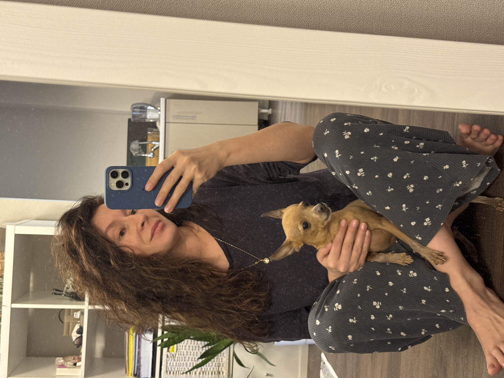 A woman with long brown curly hair is sitting on the floor, holding a small tan chihuahua, while taking a selfie in a mirror. She is wearing a dark grey top and patterned pants with tiny white flowers. Her left foot is visible to the right of her. The background shows a section of a room with a white shelving unit to the left, featuring various items. Further back, there is a white cabinet and other indistinct objects. The location is Chaikovsky, Russia.